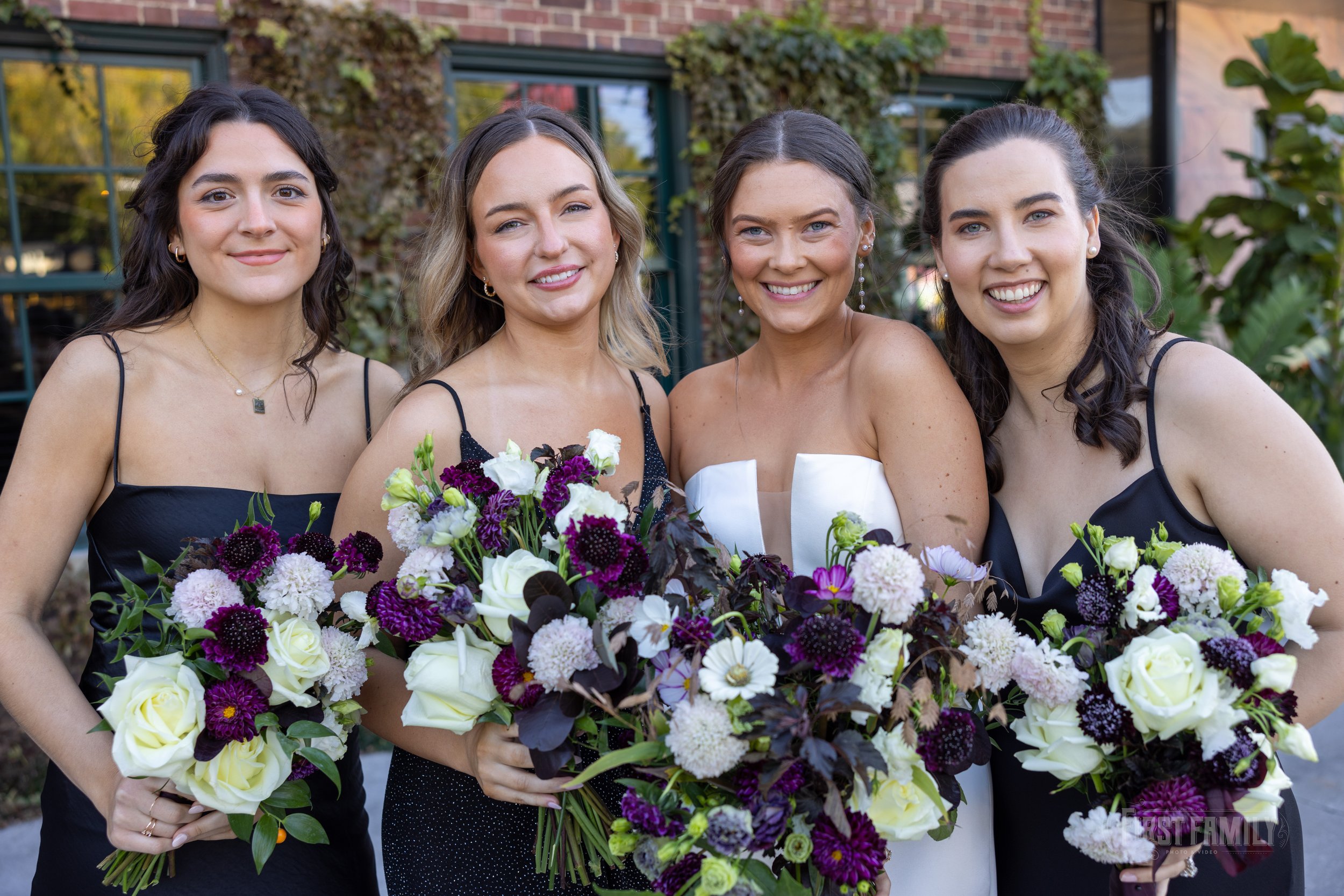 Four women in formal dresses holding large colorful bouquets of flowers, smiling outdoors in front of a building with greenery.