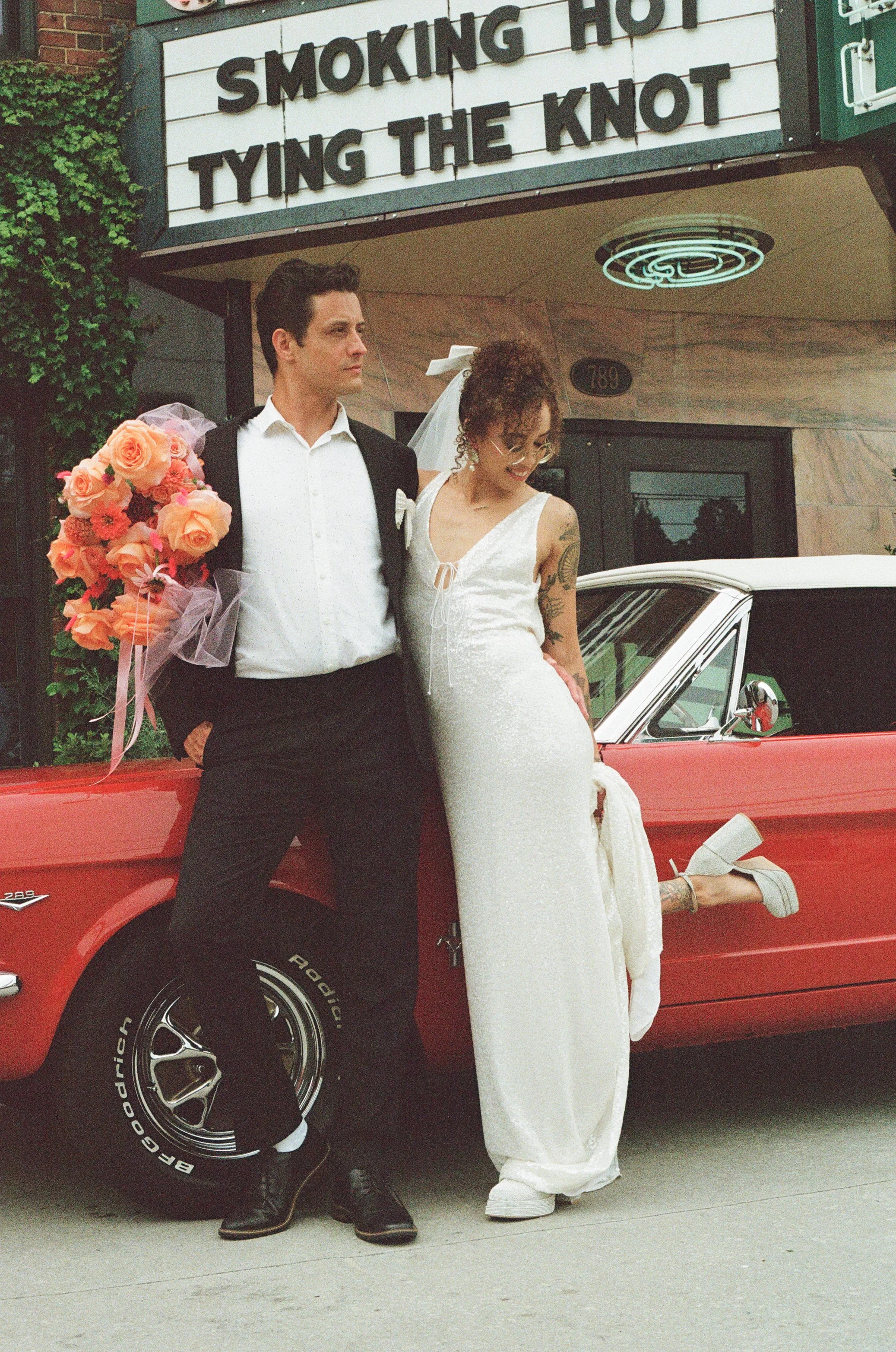 A wedding couple standing beside a red vintage car in front of a building with a sign that reads 'Smoking Hot Tying the Knot.' The groom holds a bouquet of flowers, and the bride is wearing a wedding dress and white heels.