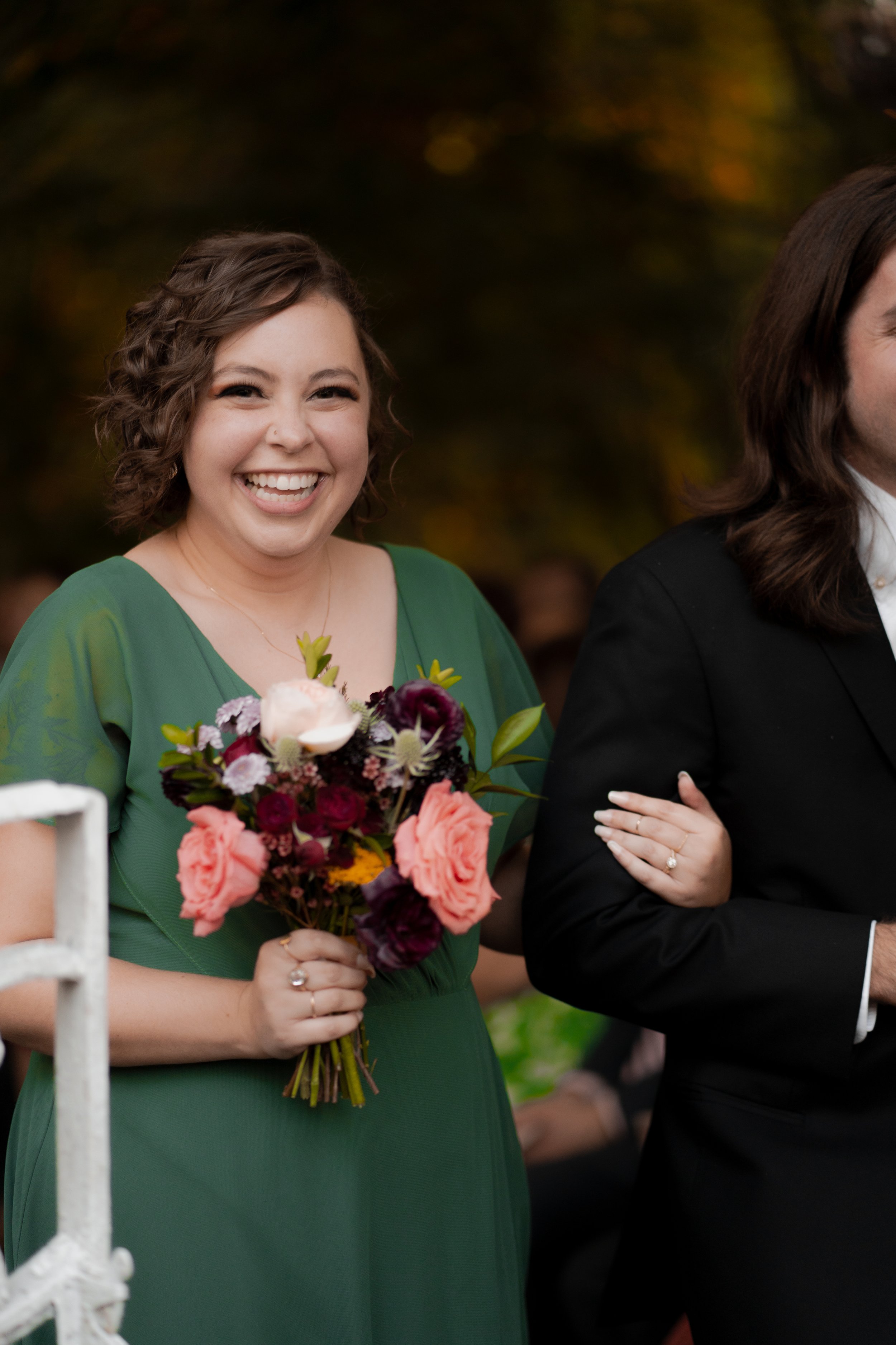 Woman in green dress smiling and holding a bouquet of flowers at an outdoor event.