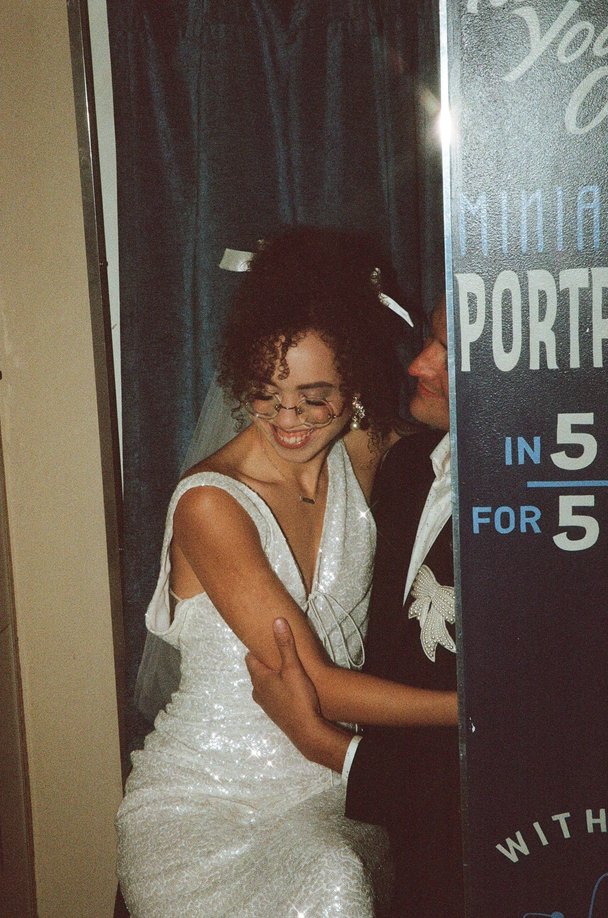 A smiling bride in a shiny white wedding dress with glasses and curly hair, standing close to a groom in a tuxedo, both pressing against a photo booth background with a playful posture.