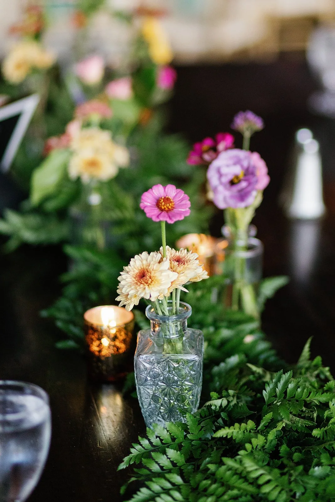 Vase with pink, purple, and cream flowers on a dark table, with a lit candle nearby and green foliage.