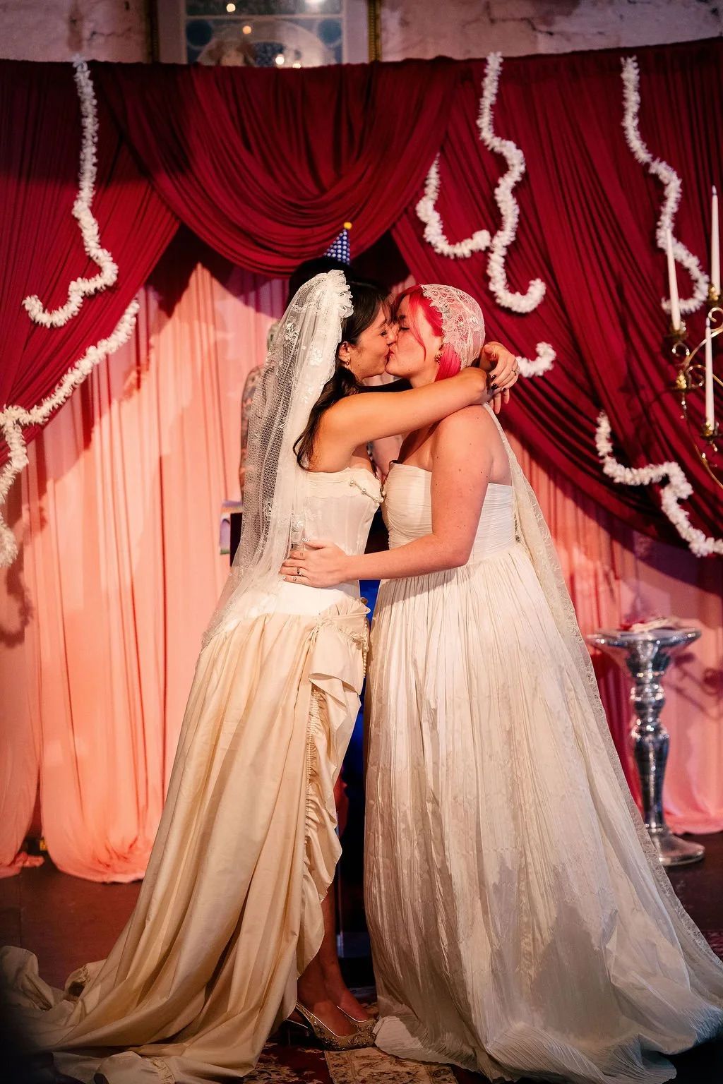 Two women wearing wedding dresses are kissing on a decorated stage with red curtains and white garlands.