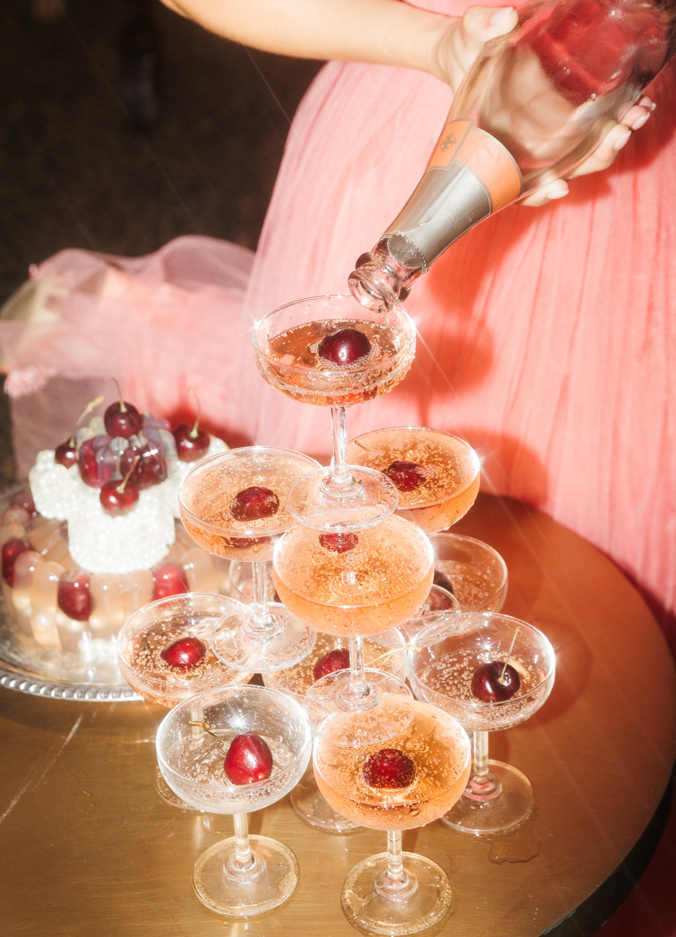 Person pouring pink sparkling drink with cherries into a champagne glass on a tiered display of similar drinks, with a cake topped with cherries in the background.