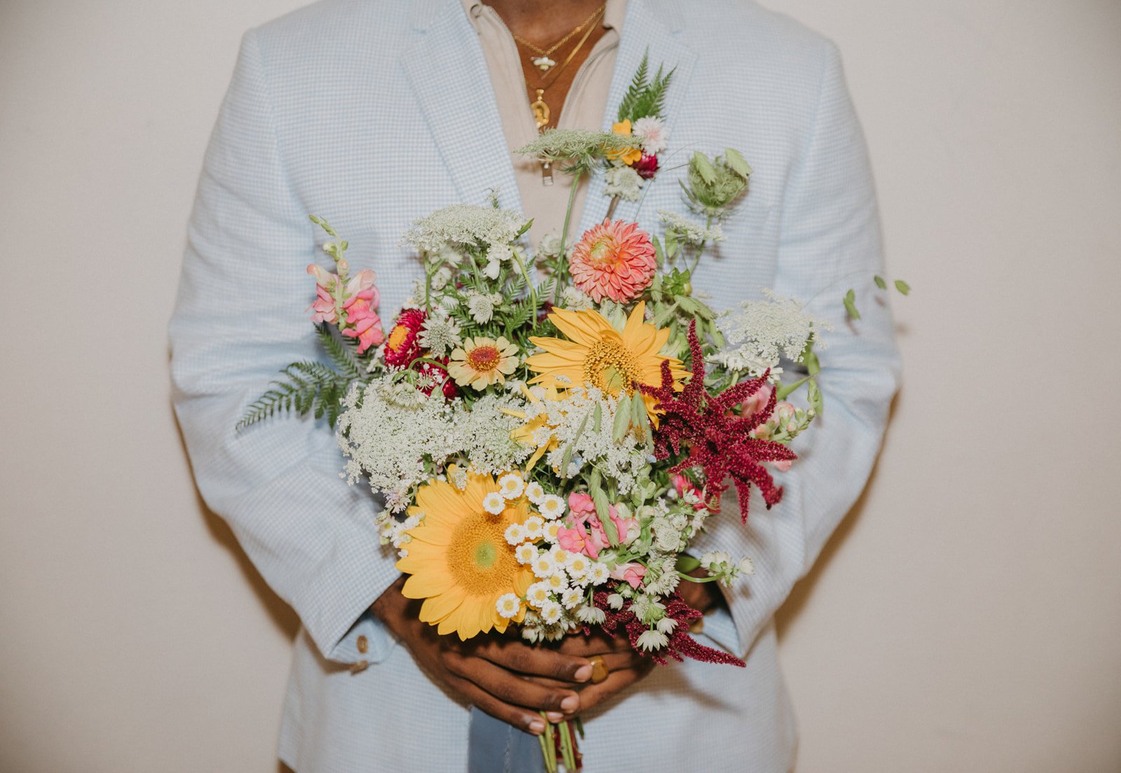 Person holding a colorful bouquet of mixed flowers including sunflowers, daisies, and pink and red blooms, wearing a light-colored blazer and gold jewelry.
