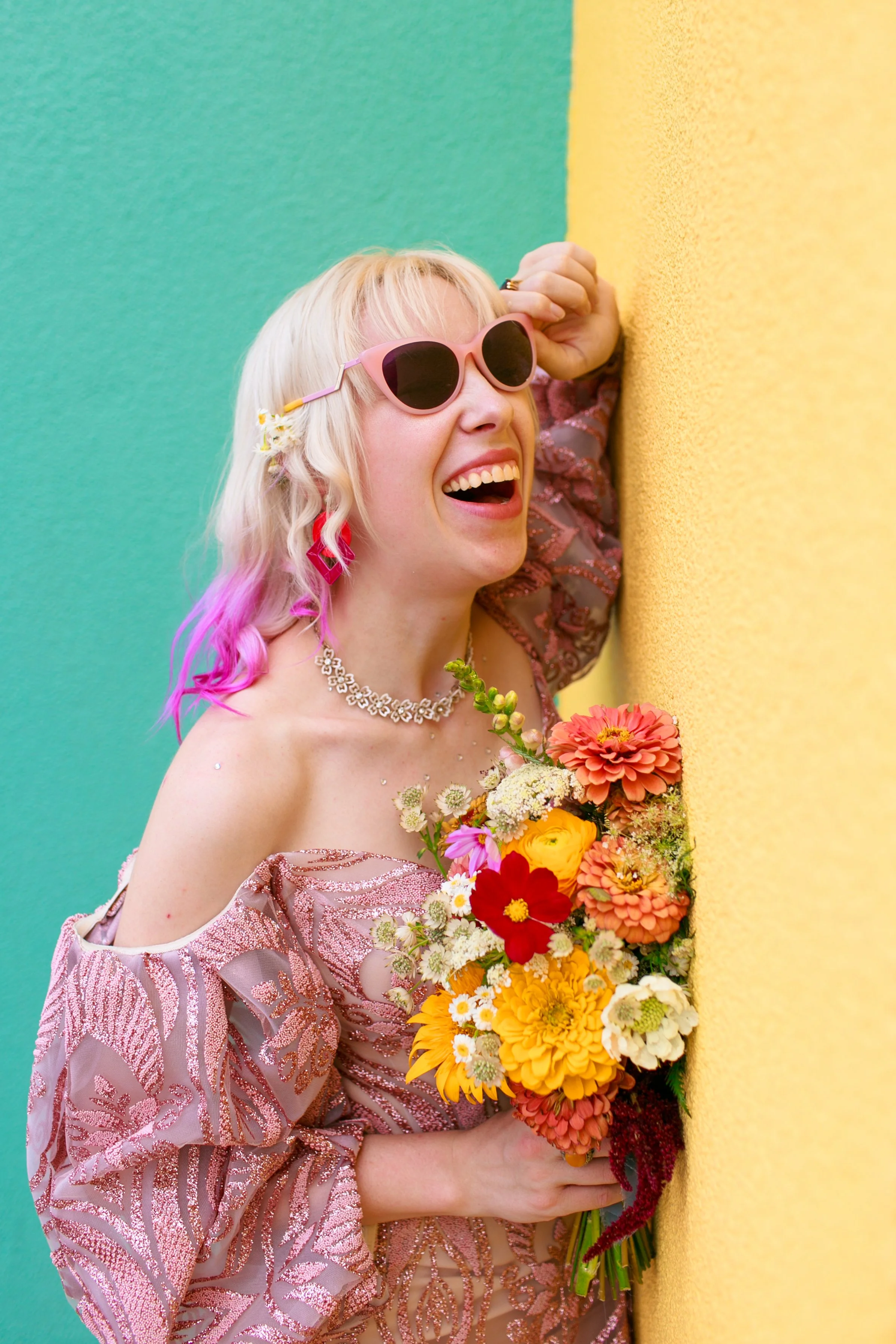 A woman with blonde hair and pink sunglasses, wearing a shiny, patterned dress, holding a colorful bouquet of flowers and laughing, positioned next to a yellow wall and a teal background.