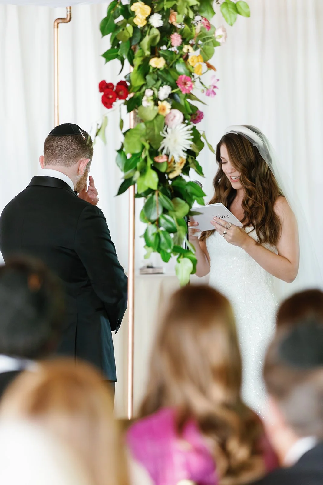 Bride and groom exchanging vows during wedding ceremony, with a floral arrangement and guests observing.