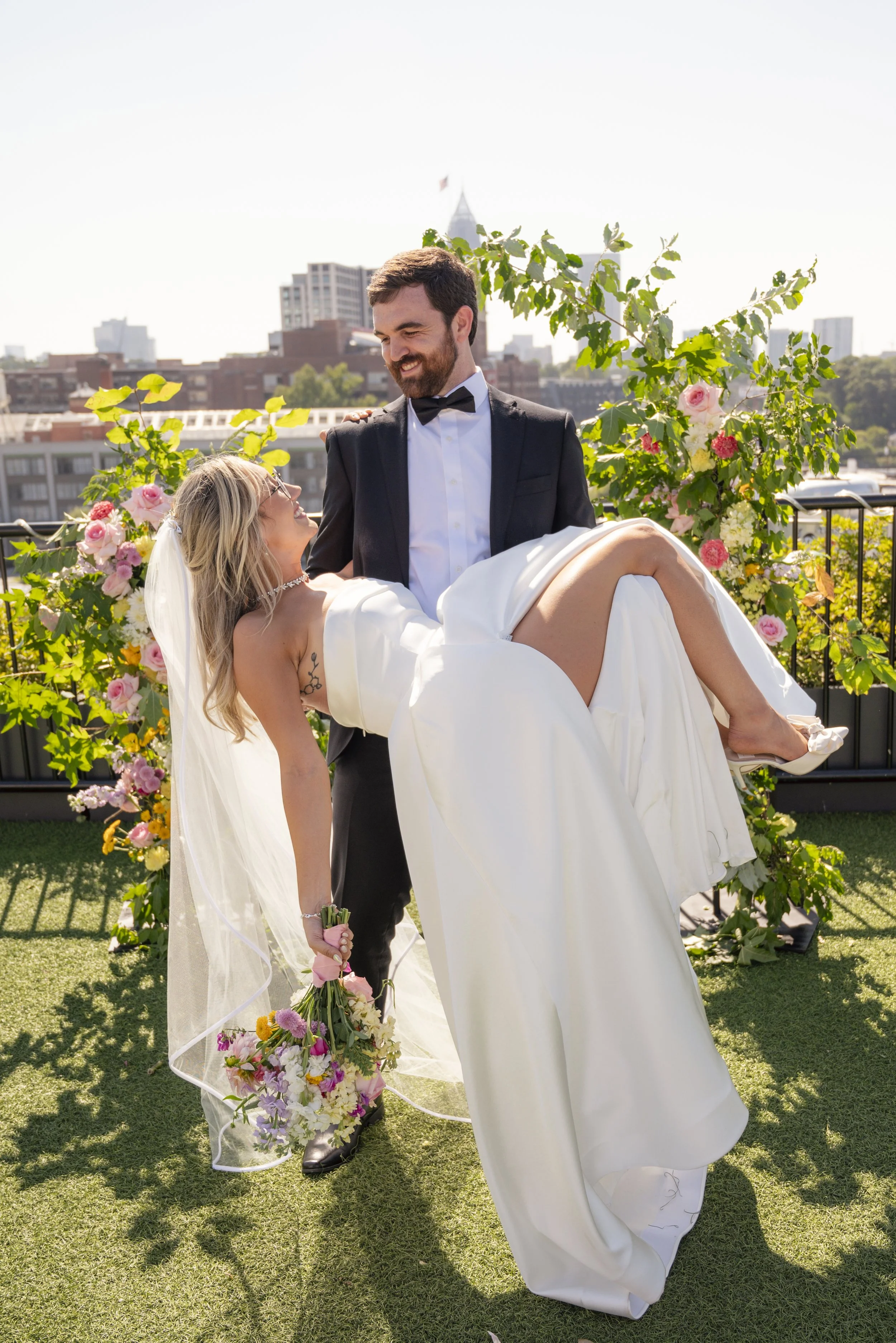 A groom in a tuxedo carries a bride in a white wedding dress and veil, smiling at her on a rooftop with city buildings and floral decorations in the background.