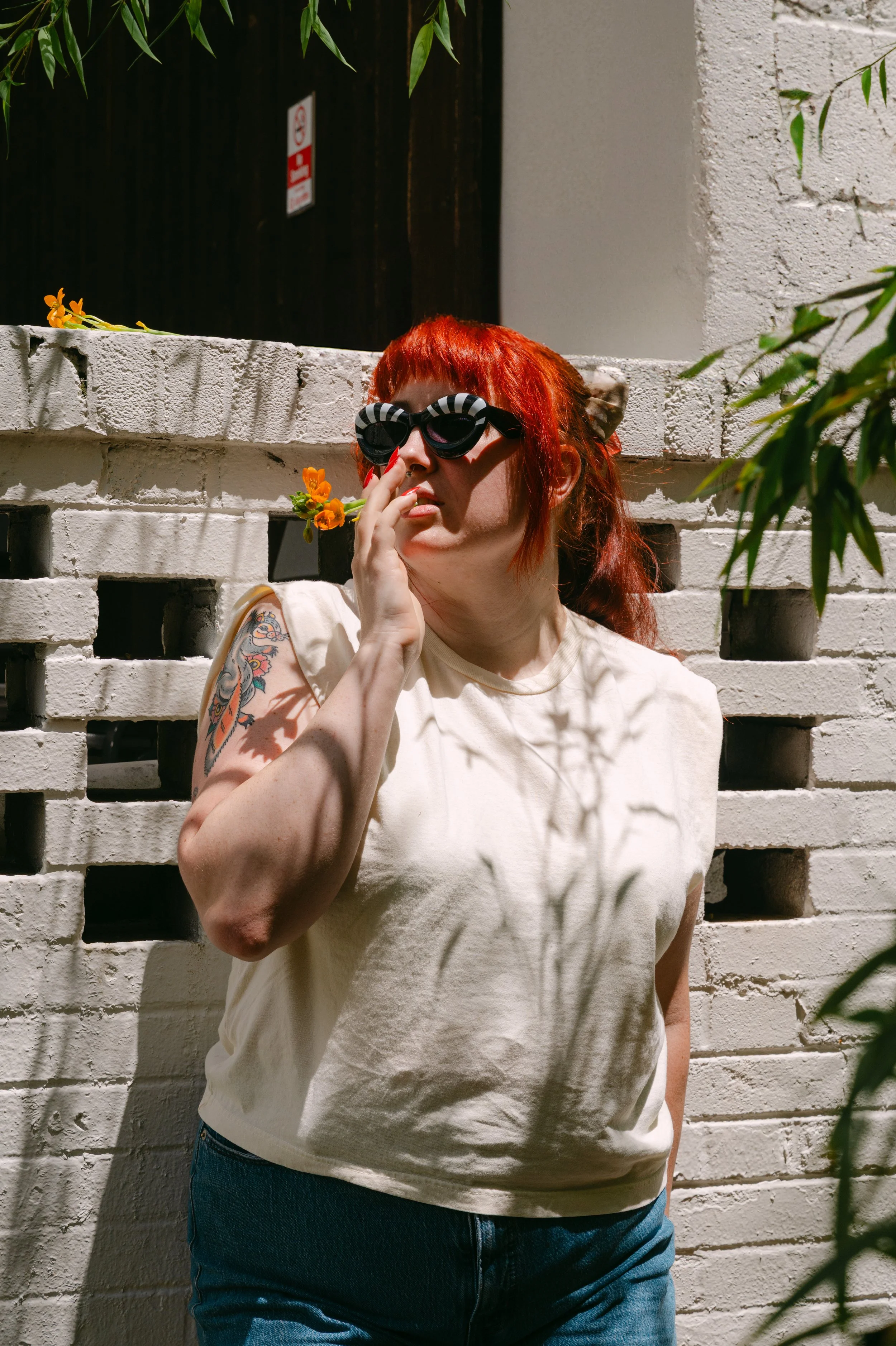 A woman with red hair and tattoos, wearing striped sunglasses and a white t-shirt, standing in front of a white brick wall with greenery and flowers.