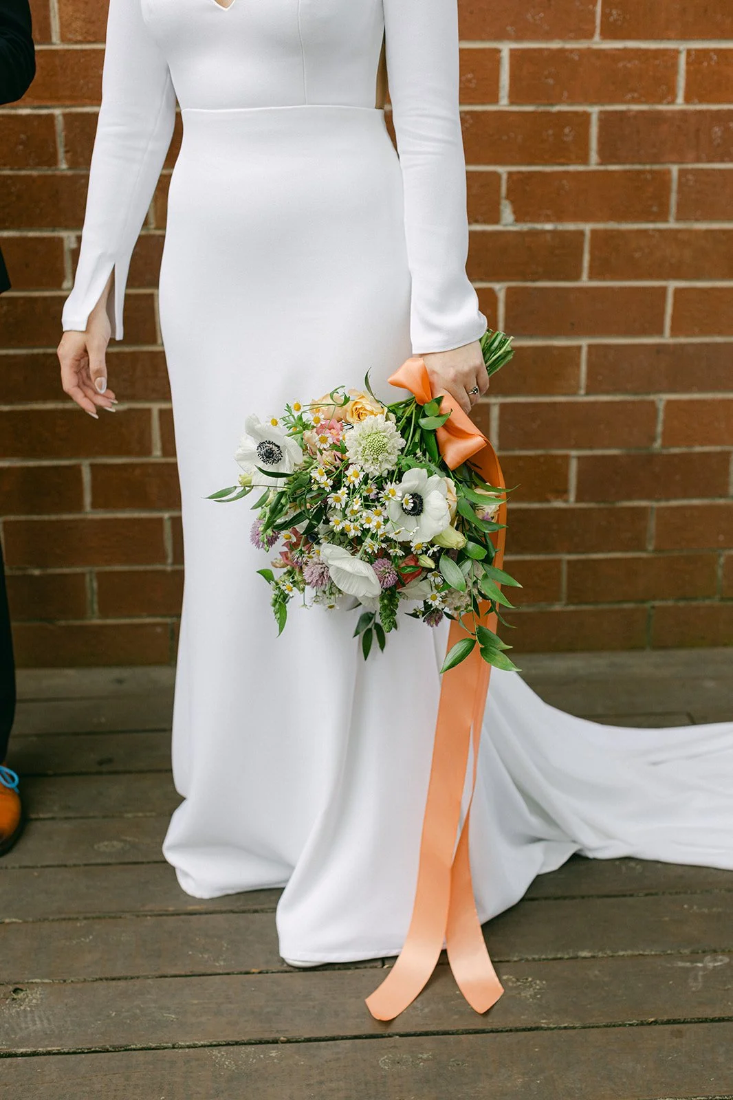 A bride in a long white wedding dress holding a bouquet of flowers with orange ribbon, standing against a brick wall.