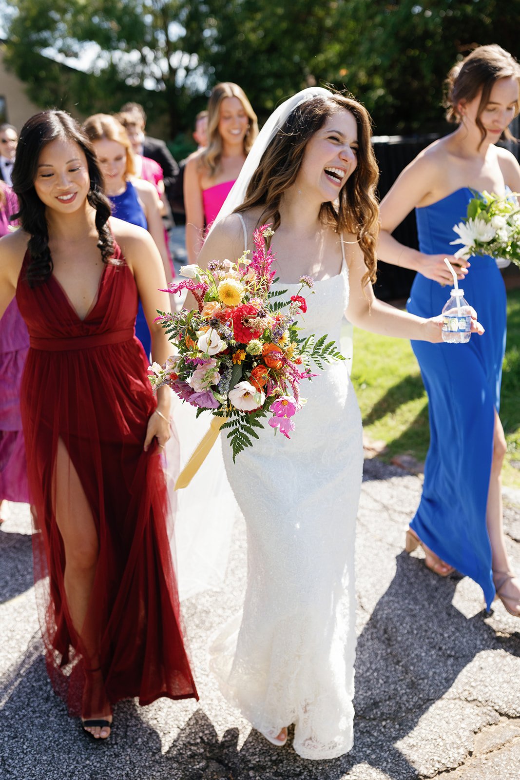 A bride in a white wedding dress carrying a colorful bouquet walks with bridesmaids in colorful dresses at an outdoor wedding ceremony.