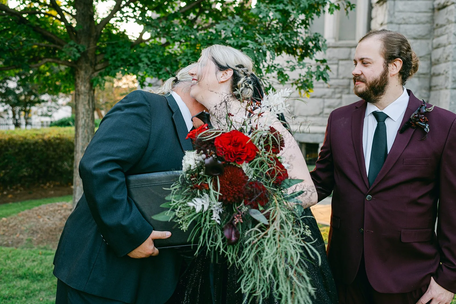 A wedding scene outdoors with two men in suits, one giving a kiss and the other smiling, with a large bouquet of red and white flowers.