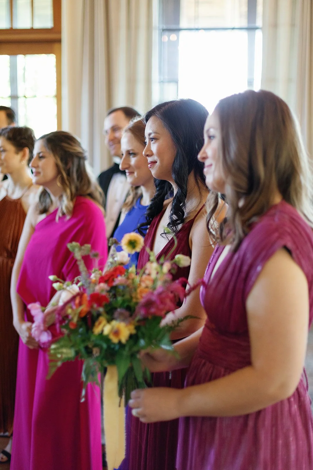 Women in colorful dresses standing in a line during a wedding ceremony, with one woman holding a bouquet of flowers.