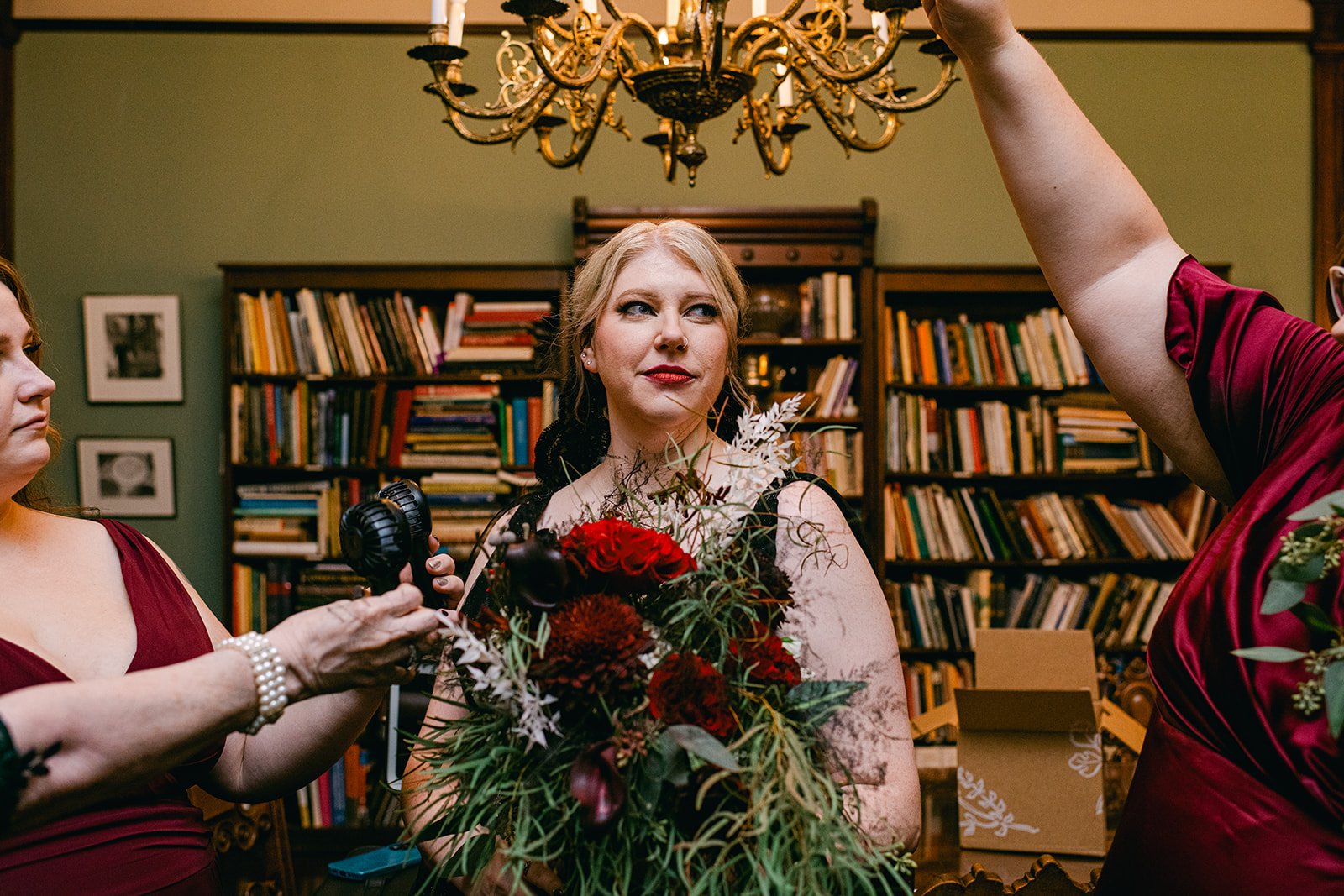A woman with blonde hair and red lipstick holding a large bouquet of red, burgundy, and white flowers. Another person, partially visible, is adjusting her hair or dress, while a third person on the left is holding a small fan. The background shows bo