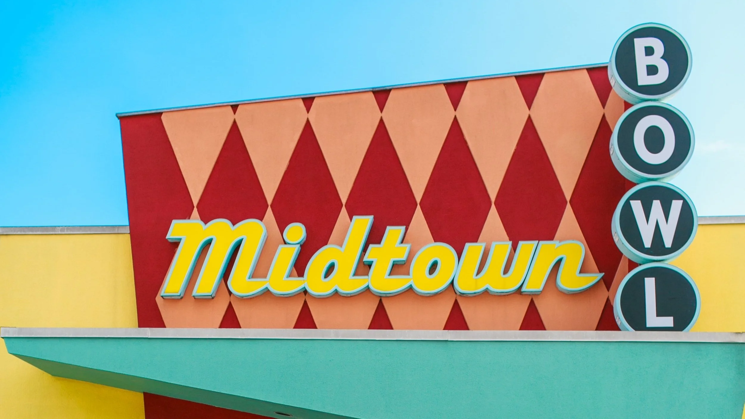 Colorful building with a red and orange diamond-patterned facade, yellow and turquoise walls, and a sign that reads 'Midtown' in yellow letters with a blue outline. To the right, there are round signs spelling out 'BOWLI' vertically.
