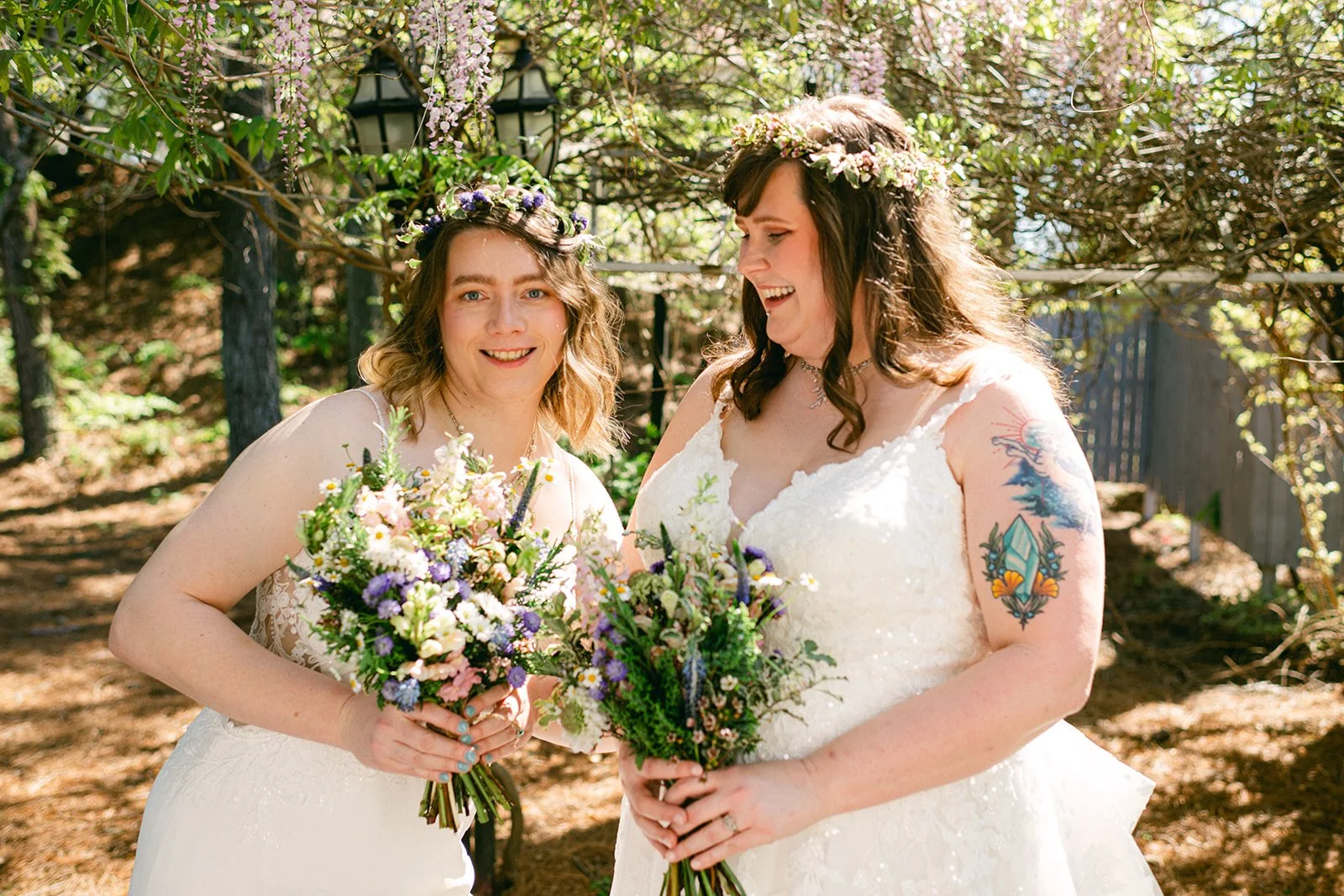 Two women in white wedding dresses with floral headbands, holding bouquets of flowers, standing outdoors among trees and flowers, smiling and looking happy.