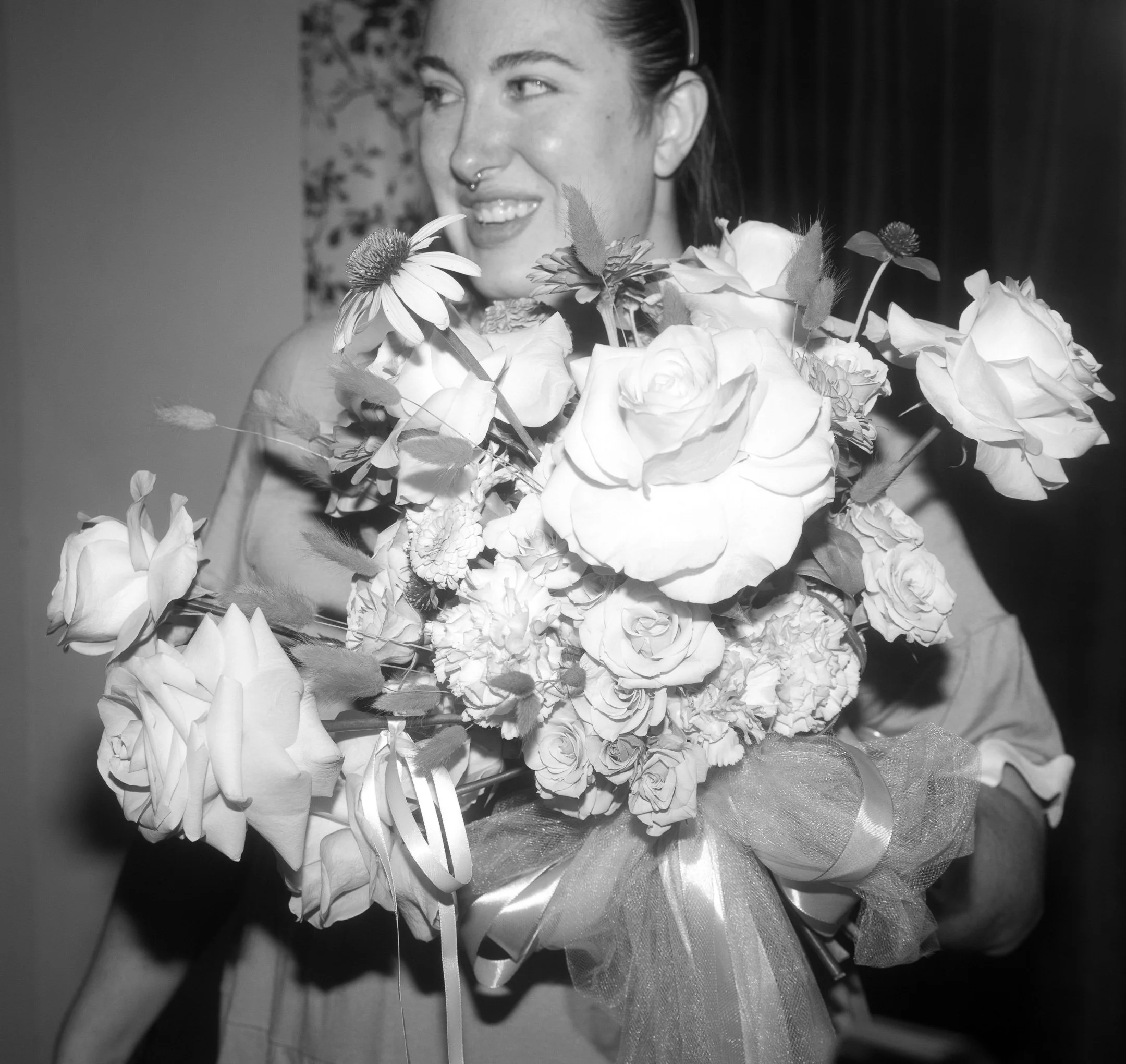 A woman with a septum piercing holding a large bouquet of flowers and looking to the side. She is wearing a hair accessory and is surrounded by various floral arrangements, in black and white.