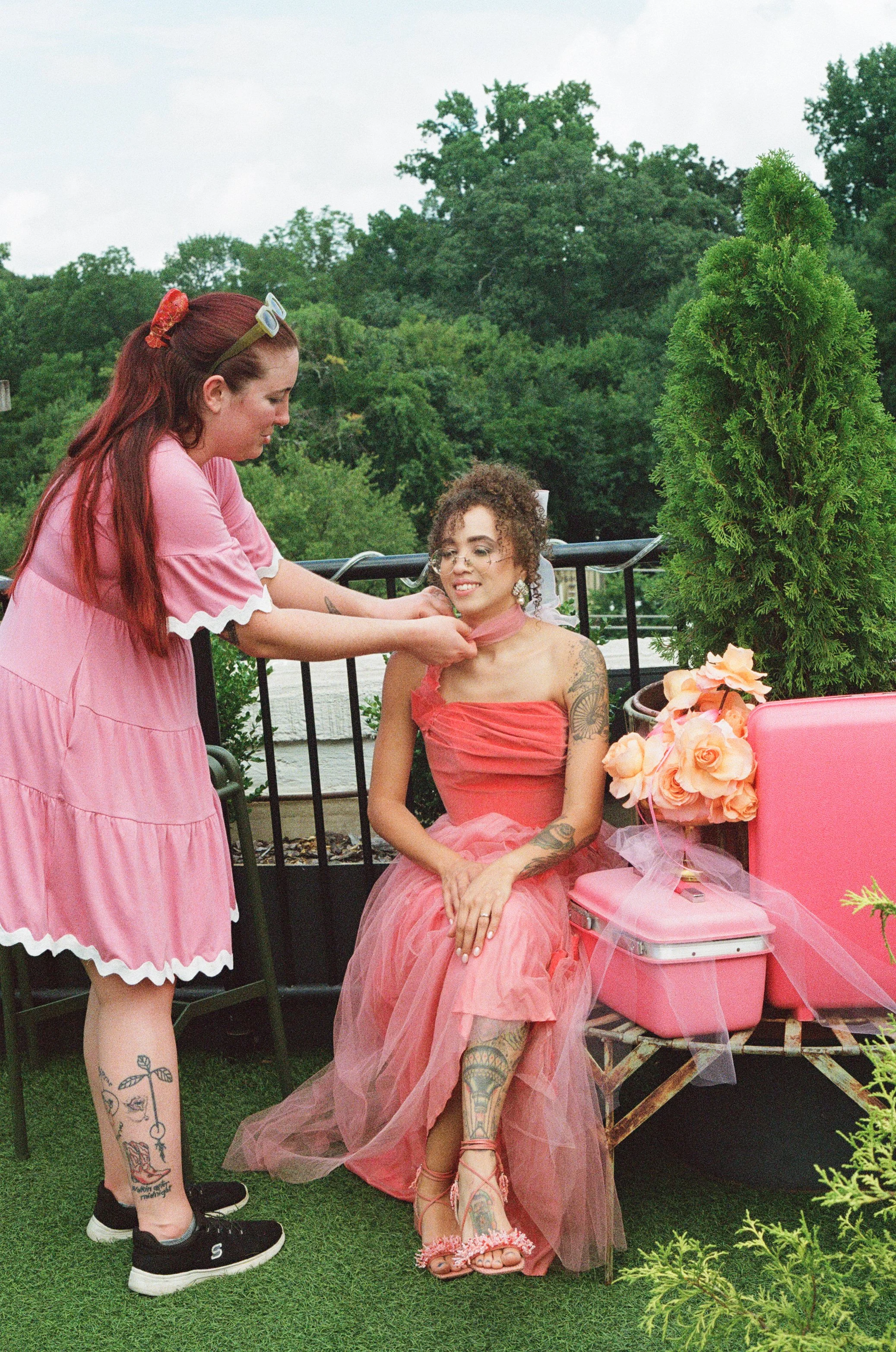A woman in a pink strapless dress is seated outdoors on a small bench, smiling as another woman in a pink dress styles her neck with a pink scarf. There are pink flowers and a pink suitcase beside her, and green trees in the background.
