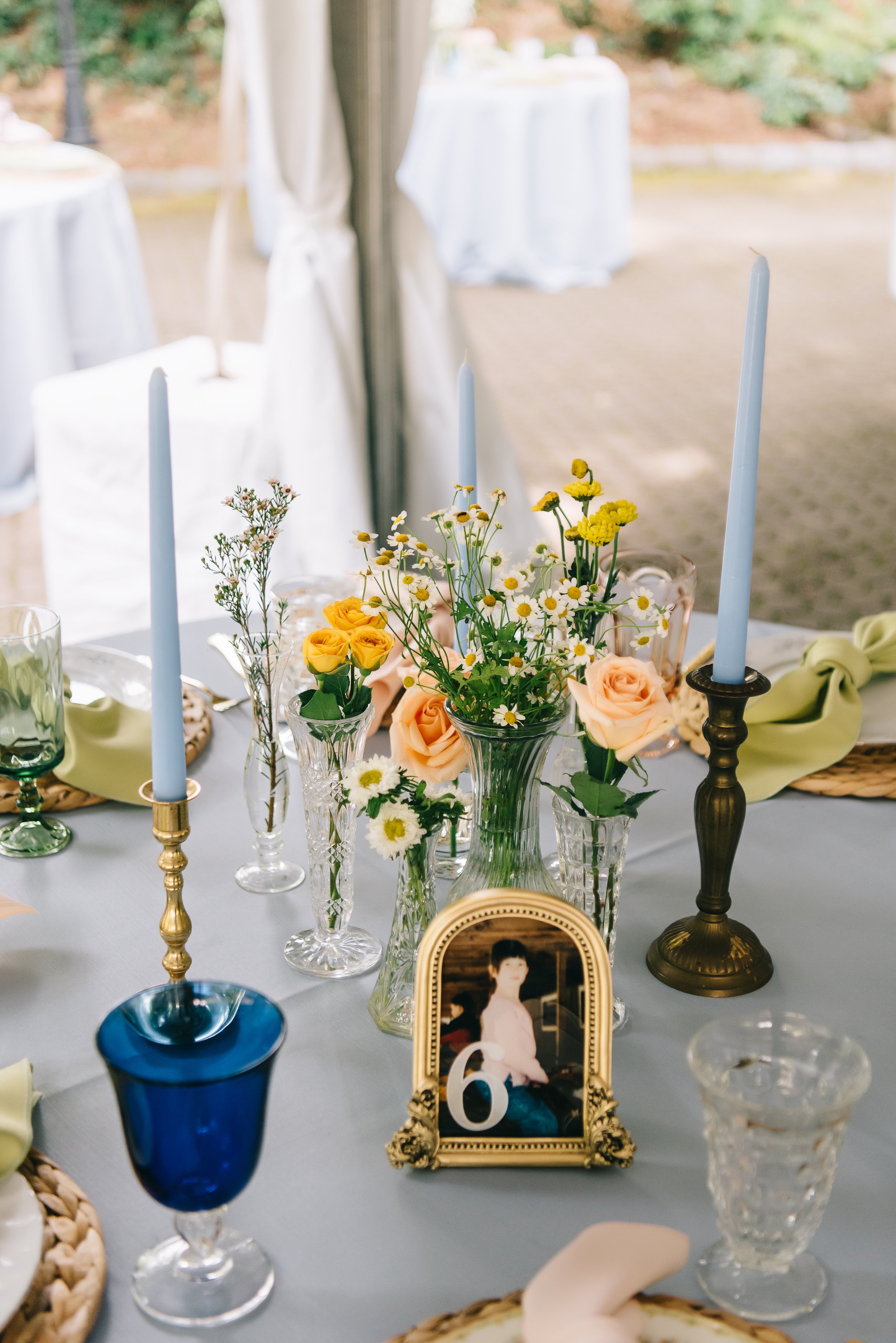Table setting with floral centerpieces, candles, glassware, a framed photograph of a woman, and a table number 6 at an outdoor event.