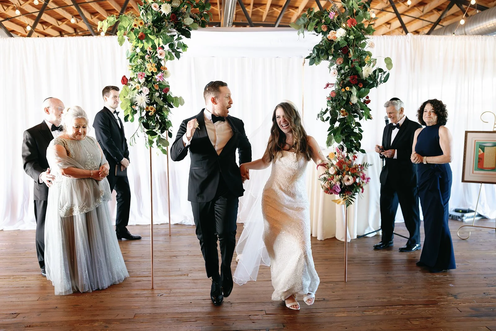 Couple dancing at wedding ceremony under floral arch with guests watching in a bright indoor venue.