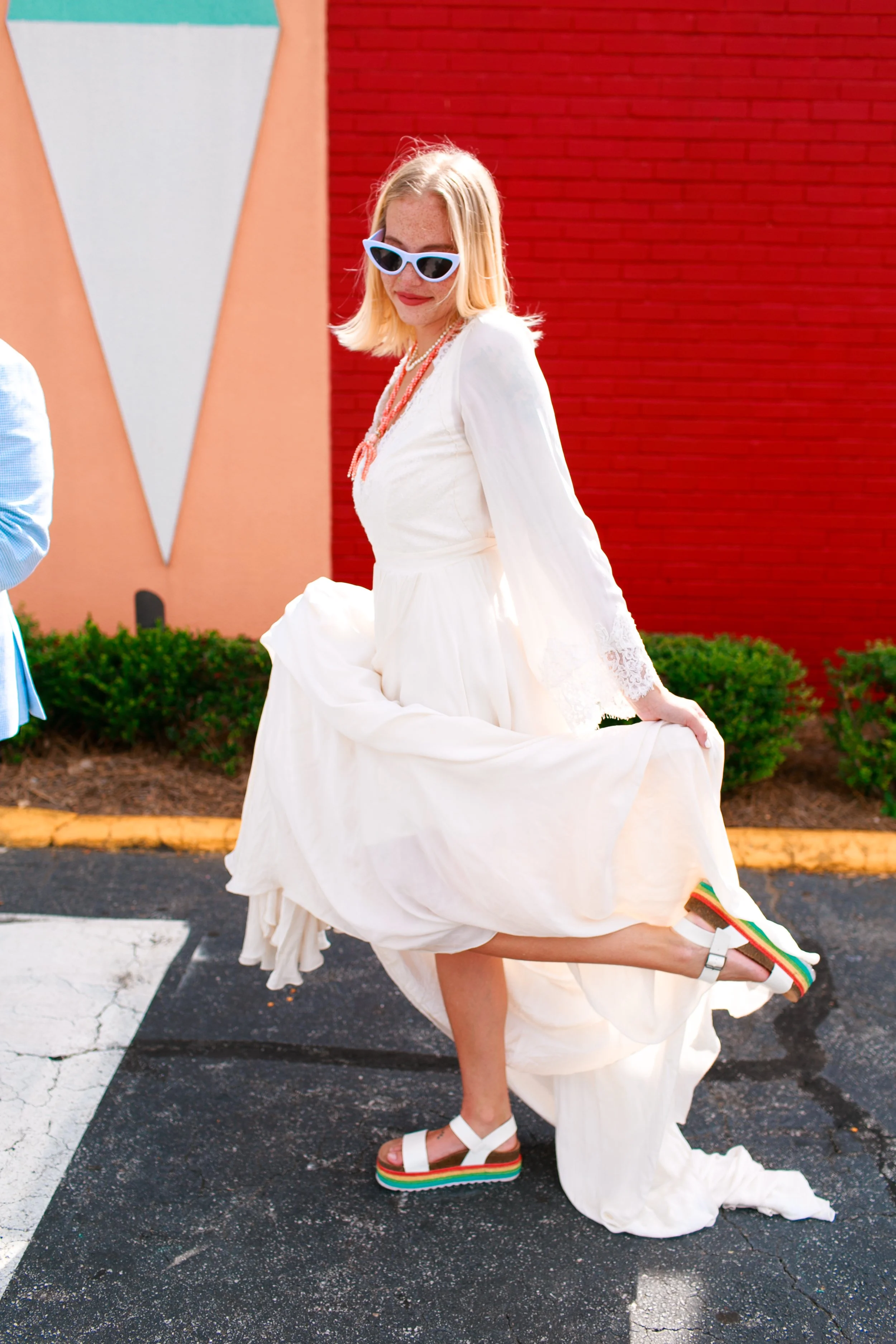 Woman wearing a white dress, blue sunglasses, and colorful rainbow sandals, lifting her dress while standing outdoors against a red brick wall with greenery.