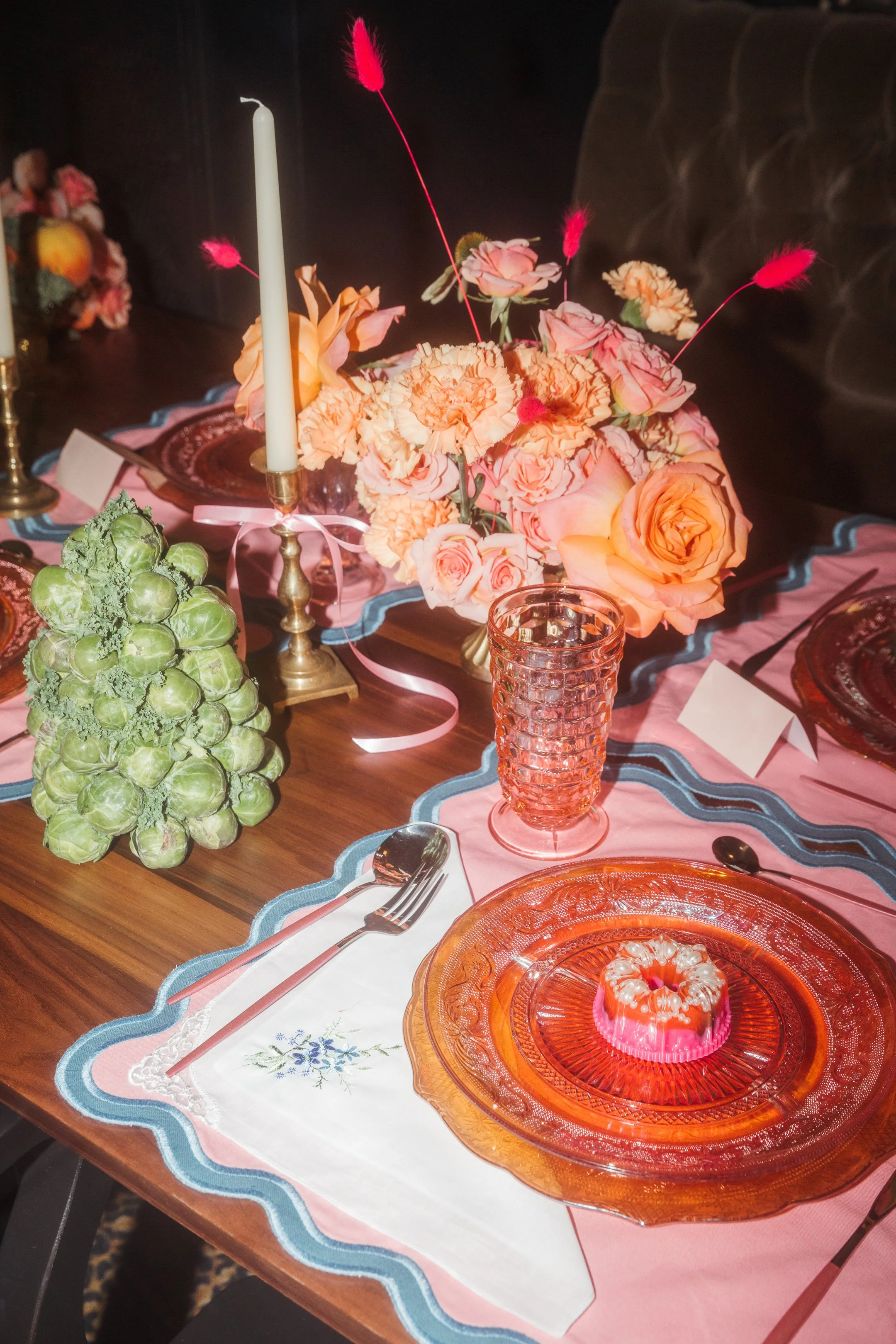 A decorated dining table featuring a centerpiece of pink and peach flowers, a brass candle holder with a white taper candle, pink and purple glassware, a white cloth napkin embroidered with blue flowers and lace, and a pink gelatin dessert on an ambe