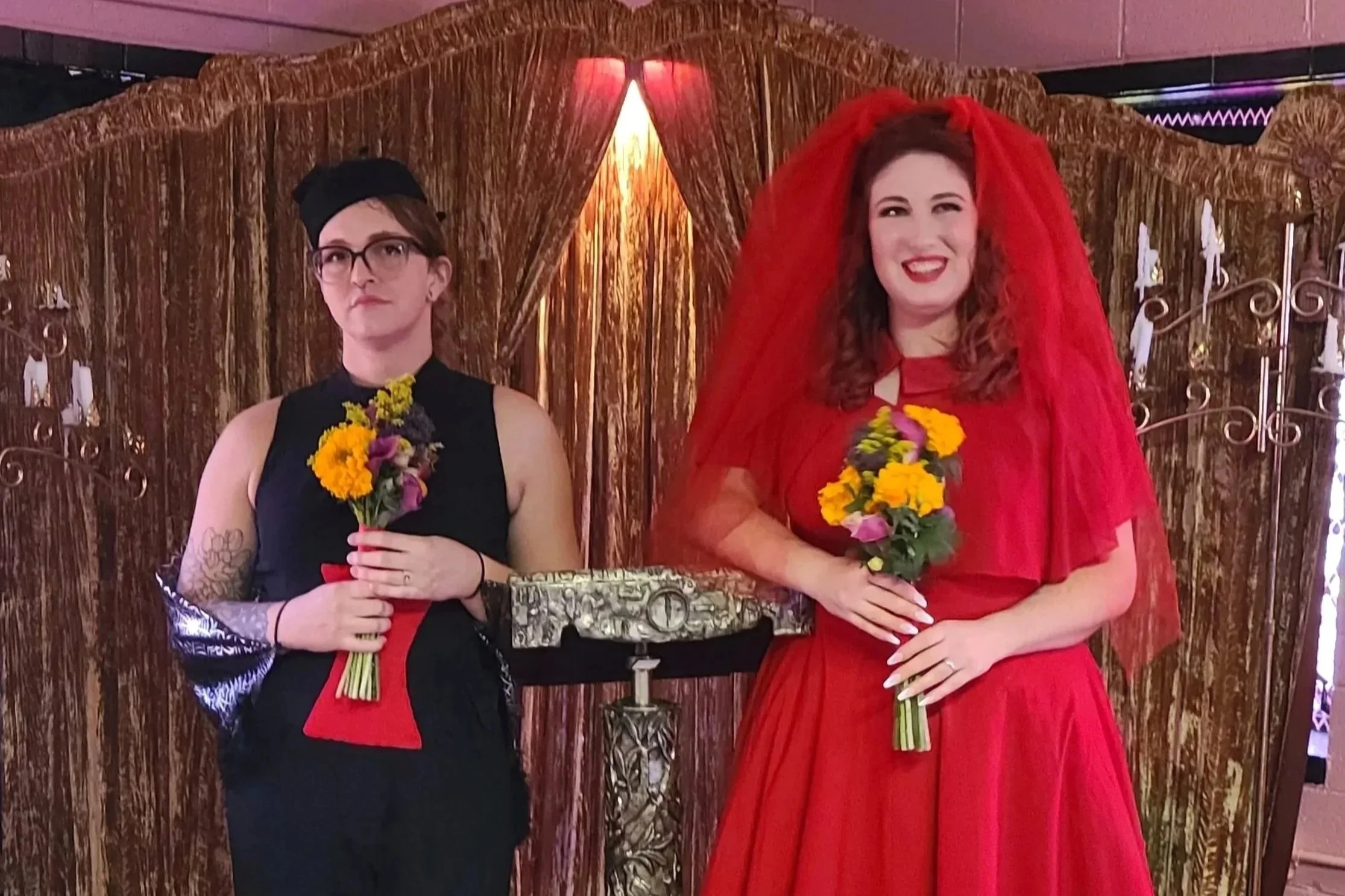 Two women standing together, each holding a bouquet of flowers, in front of a gold curtain backdrop. The woman on the left is dressed in black and red, wearing glasses and a hat. The woman on the right is dressed in a red outfit with a red veil, smiling.