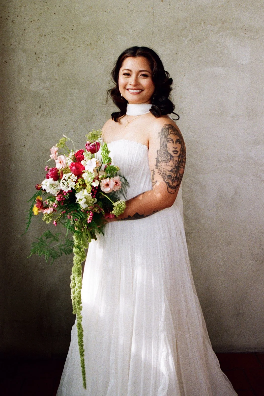 A young woman in a white wedding dress holding a colorful bouquet of flowers, smiling, standing against a plain wall background.