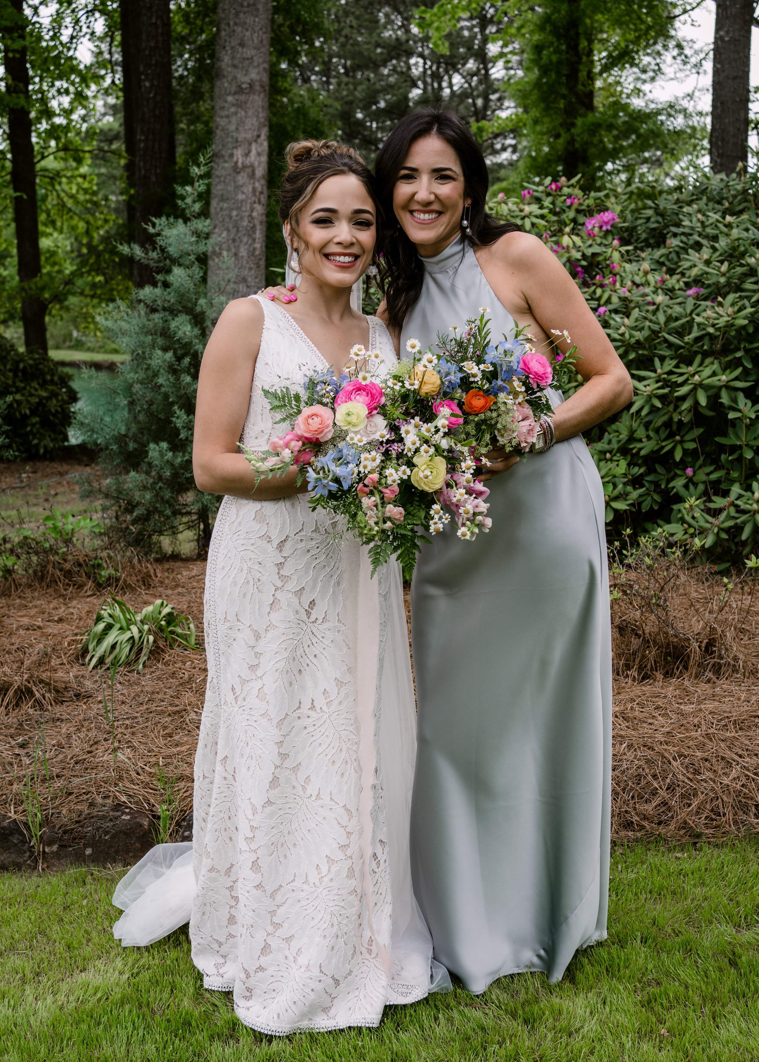 Two women in wedding attire standing outdoors amid trees and bushes, holding bouquets of colorful flowers, smiling for a photo.