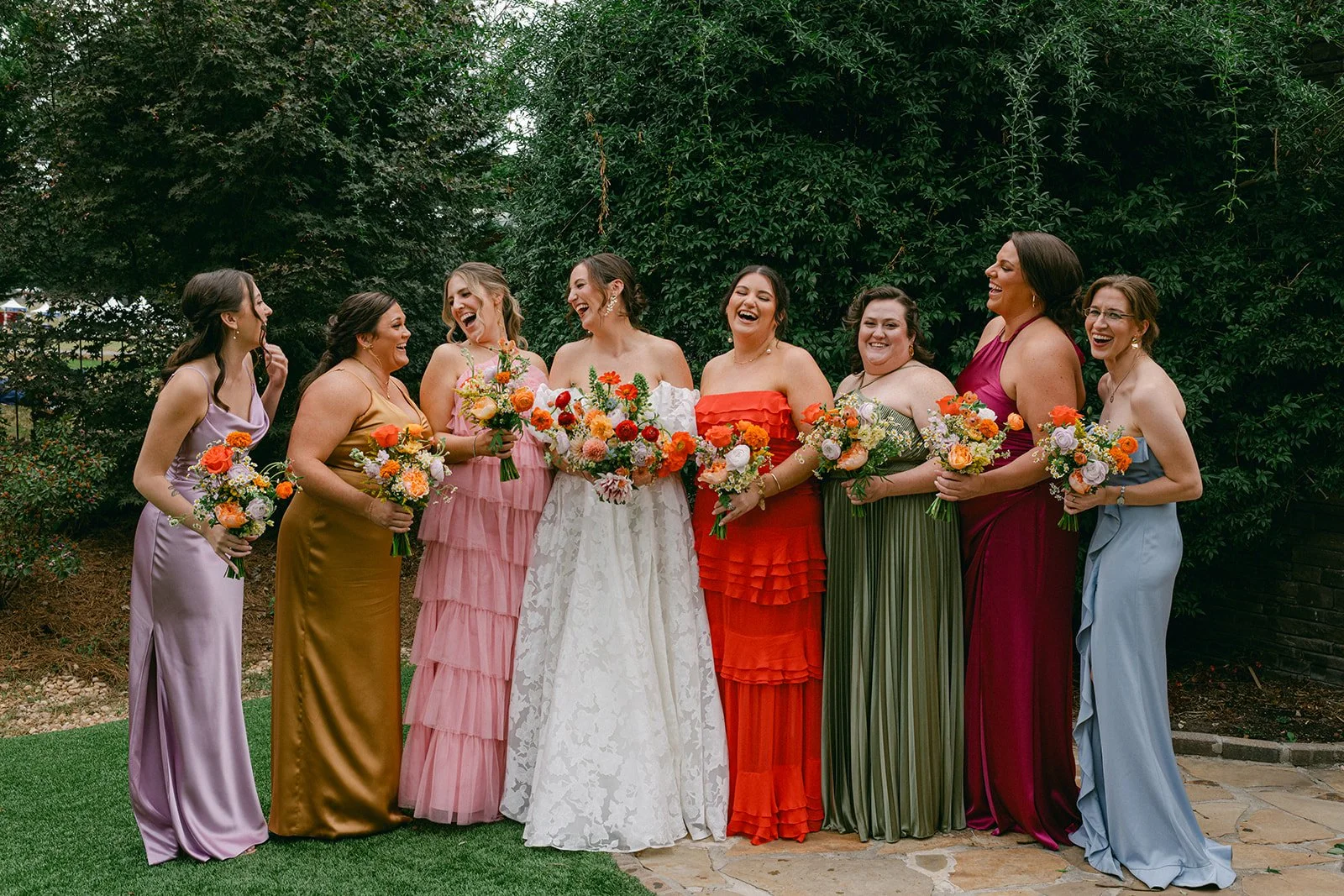Group of women dressed in colorful dresses, holding bouquets, standing outdoors in front of green bushes, celebrating a wedding.