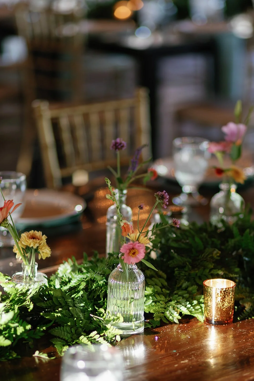 Table decorated with small glass vases of colorful flowers, green foliage, and a gold candle holder.