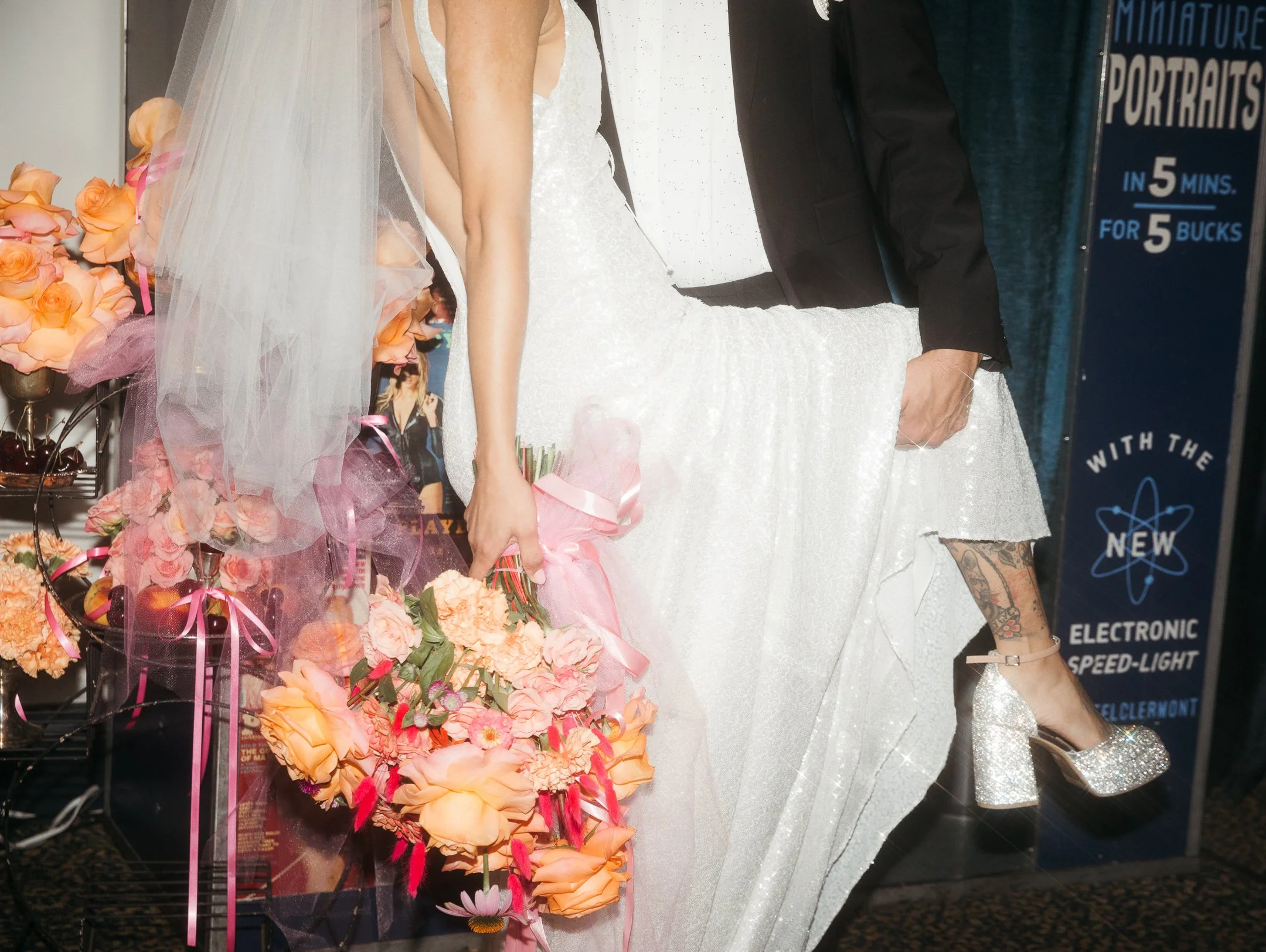 A bride holding a bouquet of pink and peach flowers, wearing a white wedding gown and veil, standing next to a man in a black suit with tattoos visible on his leg. They are at a photo booth.