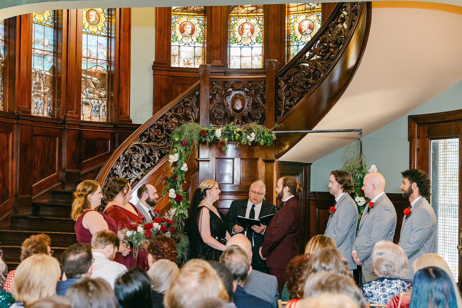 Wedding ceremony with bride and groom exchanging vows in front of officiant, with wedding party members standing on either side, inside a historic venue with stained glass windows and wooden staircase decorated with flowers.