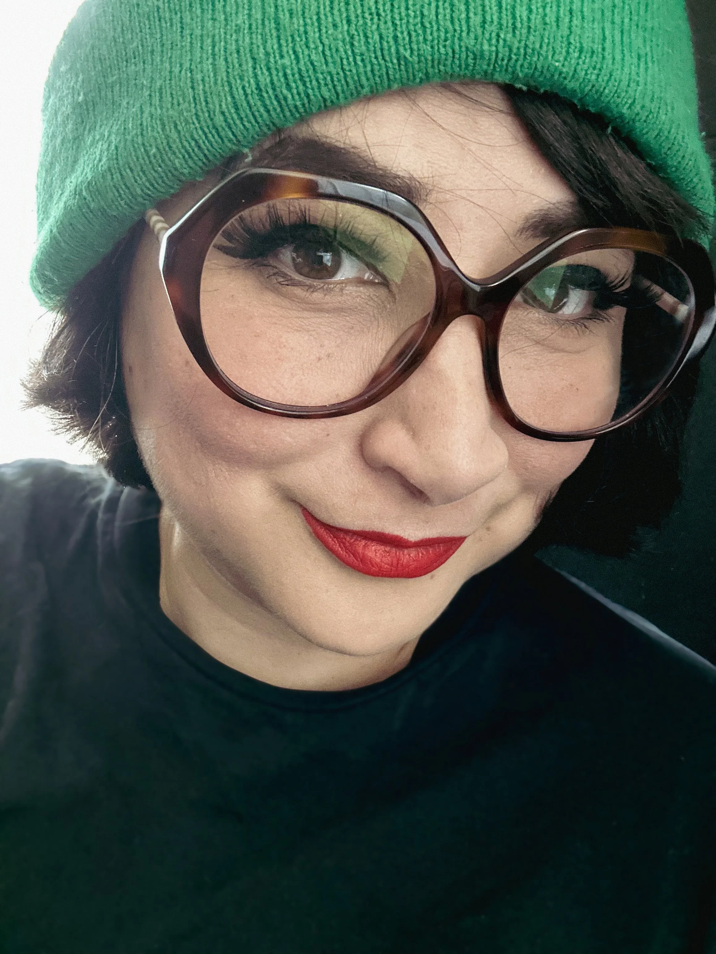 A woman with short dark hair, wearing a green beanie, large tortoiseshell glasses, red lipstick, and a black shirt.