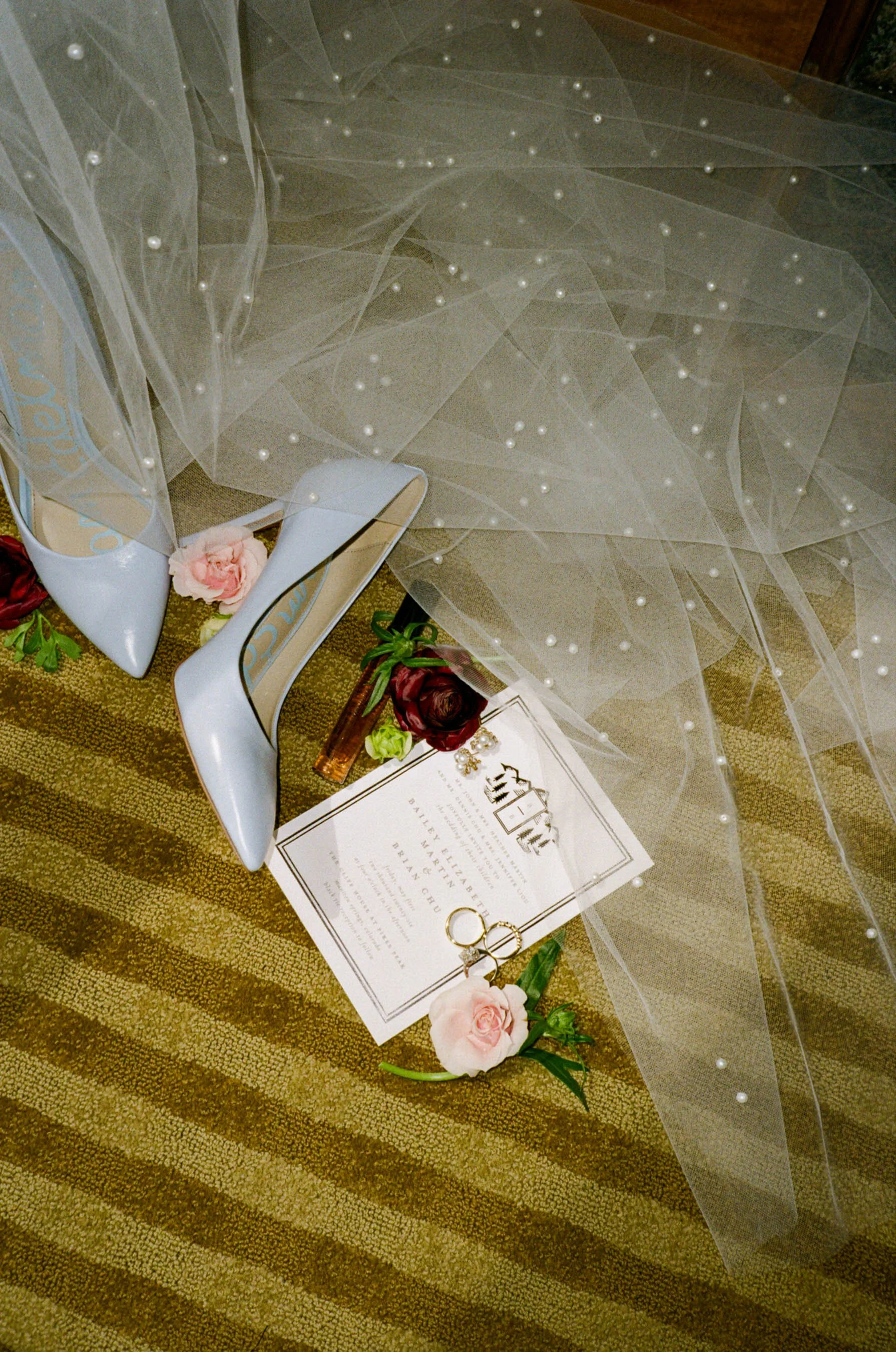 Weddings shoes, flowers, wedding rings, and a wedding invitation on a brown carpet.