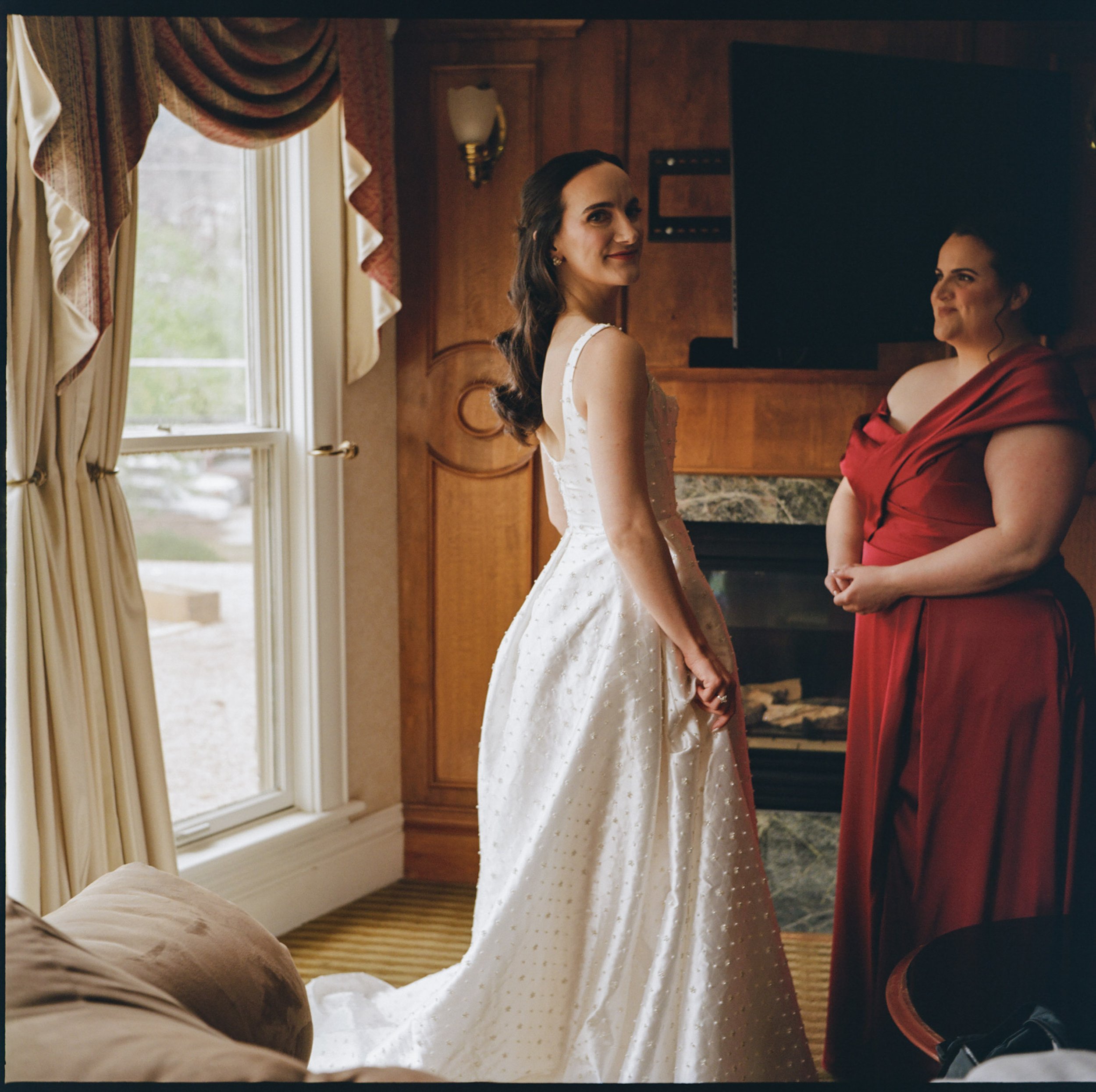 Two women standing face-to-face in a warmly lit room with wooden walls, near a window with curtains. The woman on the left is wearing a white dress with small dots, and the woman on the right is wearing a red dress. They are holding hands, and one wo