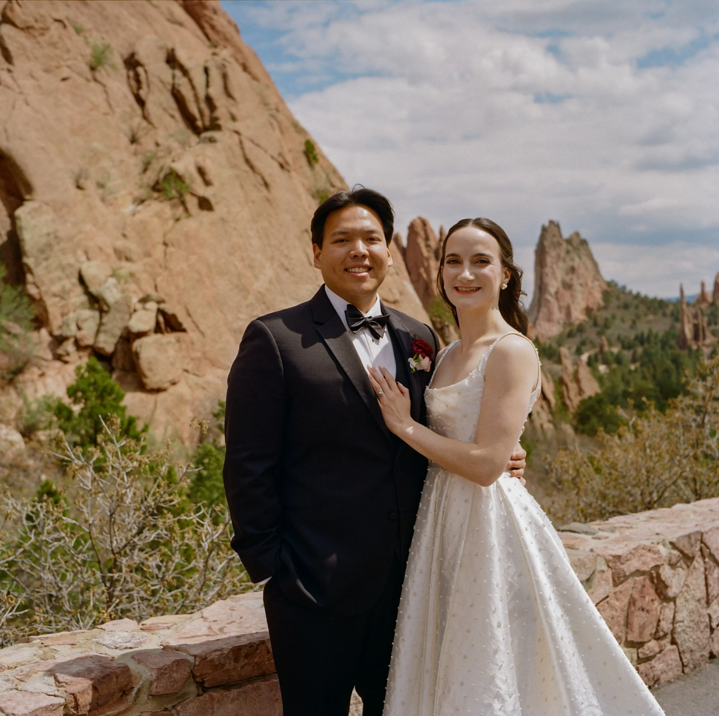 A wedding couple dressed in formal attire posing outdoors with rocky formations and a cloudy sky in the background.
