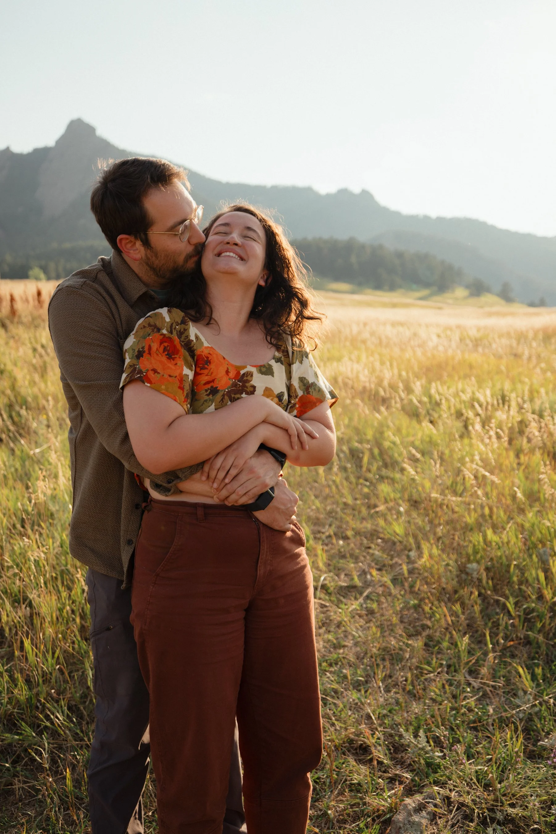 A couple stands in a field of tall grass, with mountains in the background, enjoying a sunny day. The man is kissing the woman on the cheek as she smiles with her eyes closed.