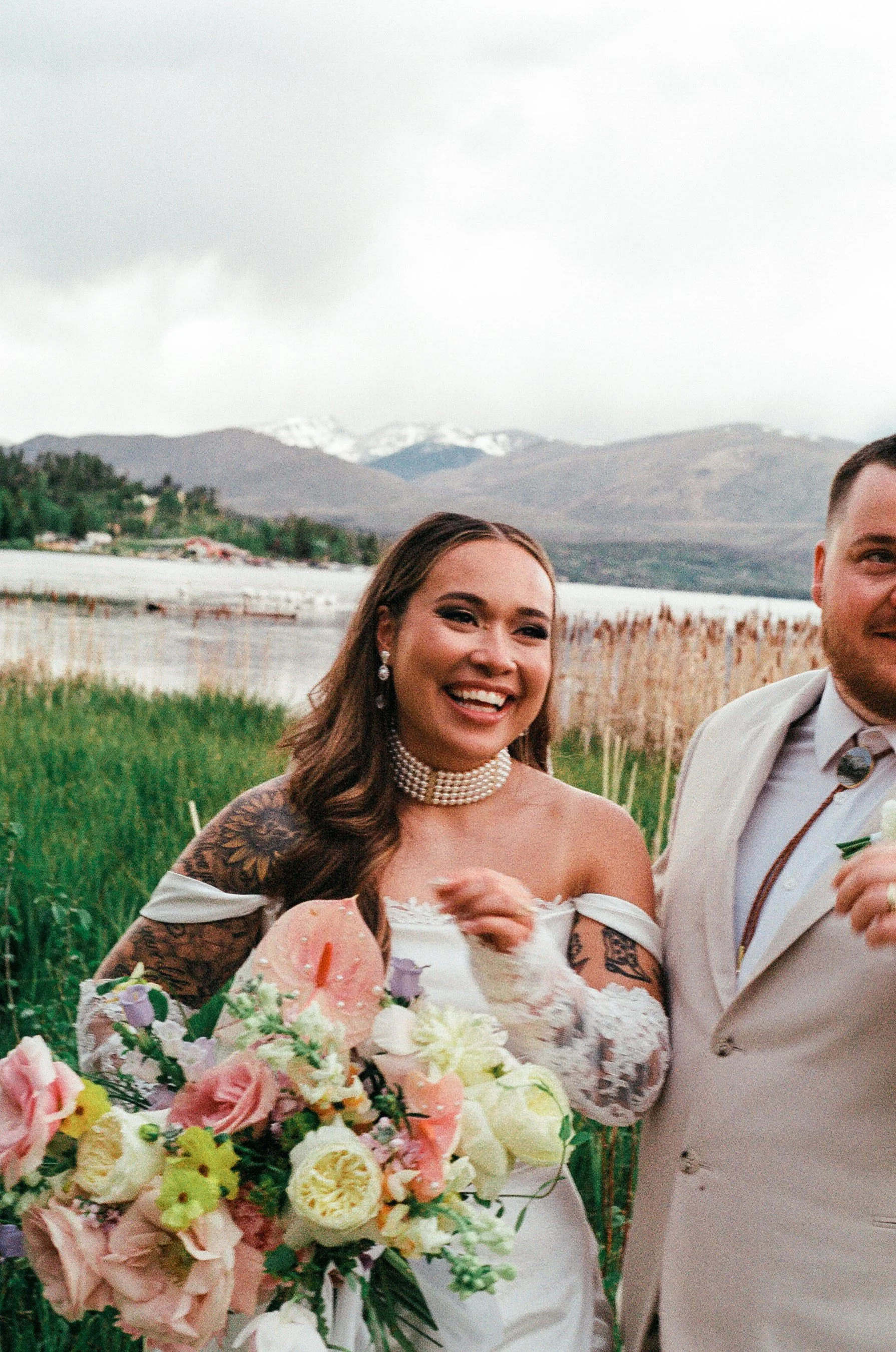 Bride smiling holding a bouquet of flowers standing beside groom outdoors with lake and mountains in background.