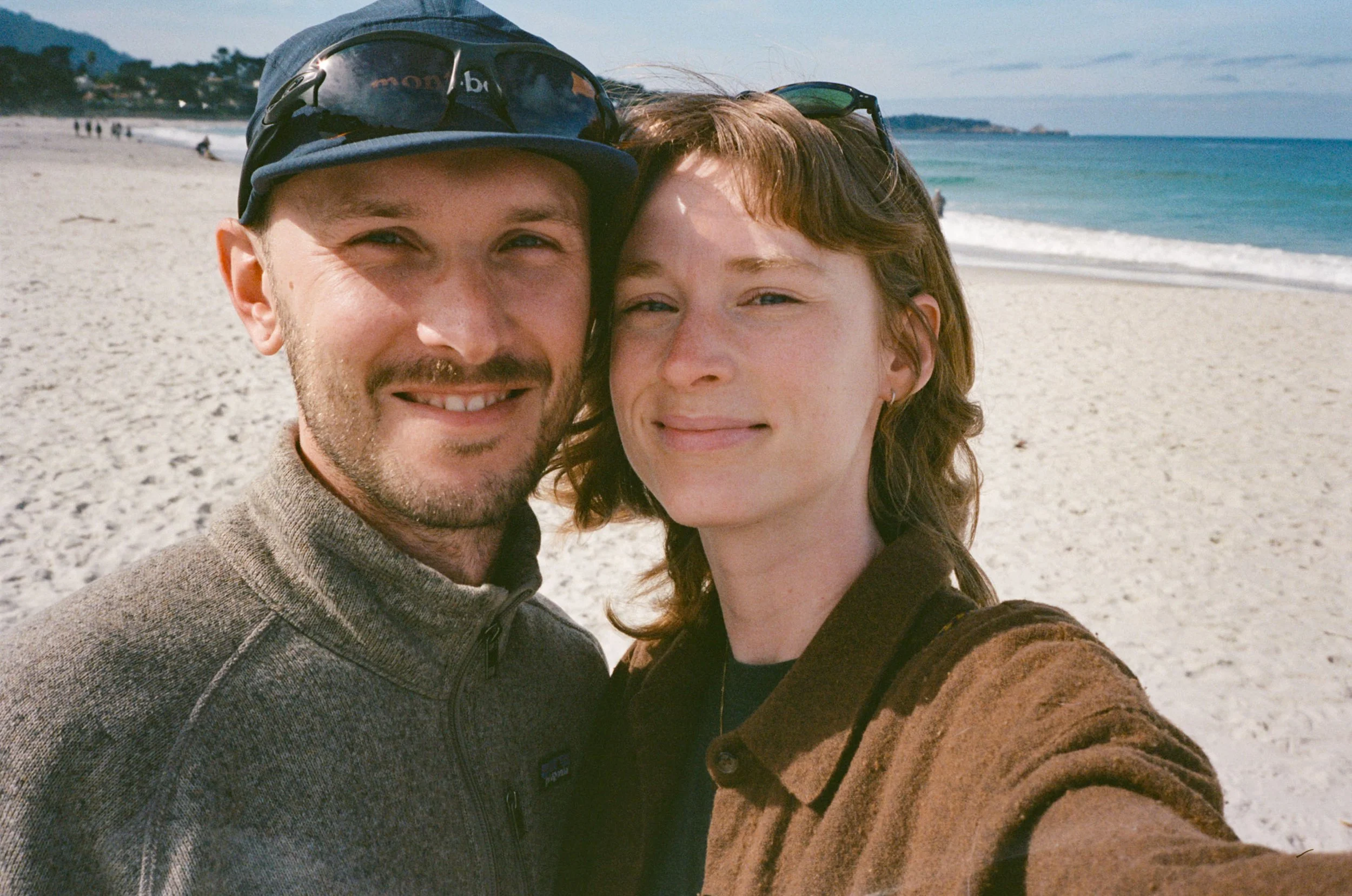 A selfie of a man and woman on a beach, smiling with the ocean and shoreline in the background.
