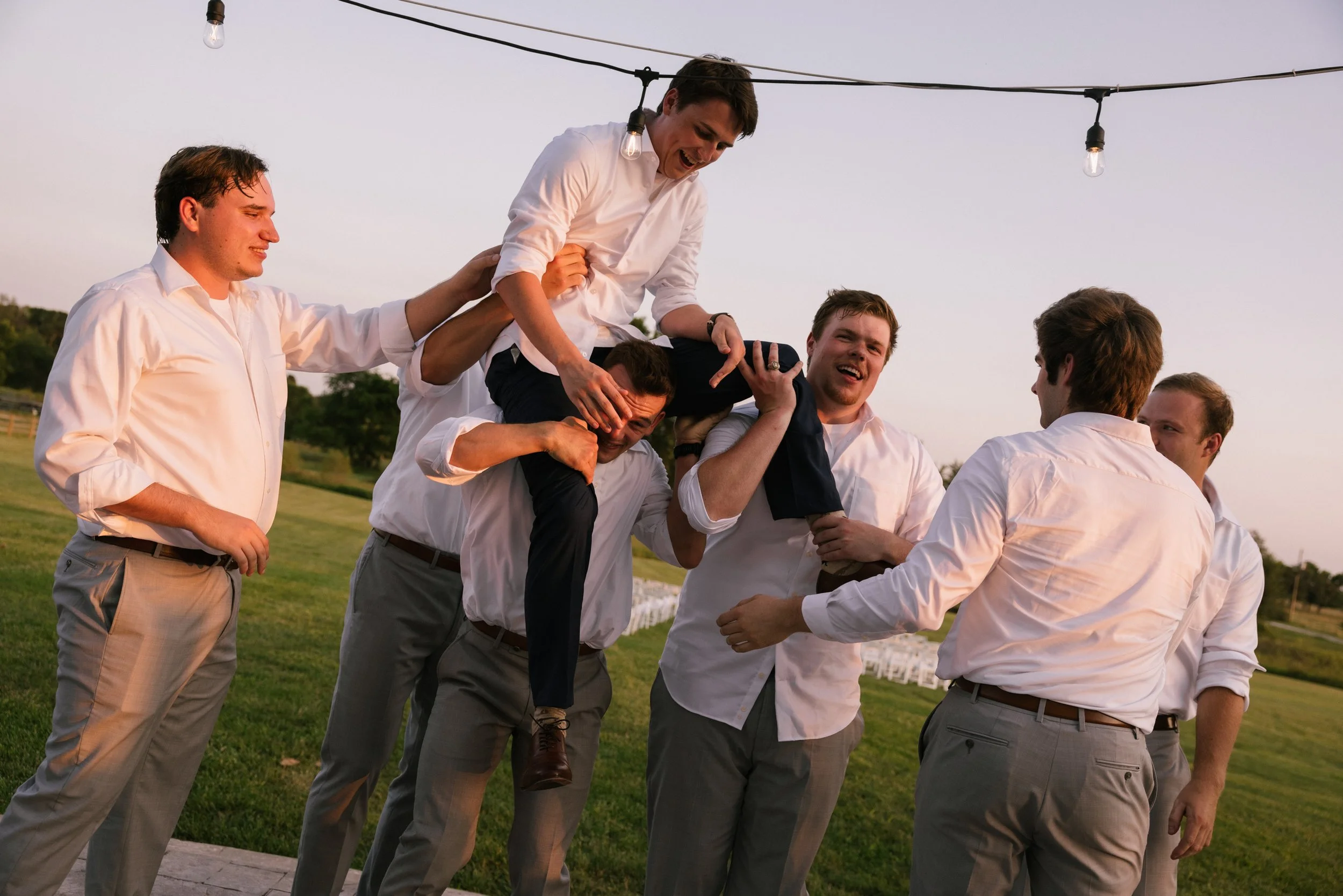 Group of men in white shirts and gray pants participating in a celebratory toast outdoors with string lights.
