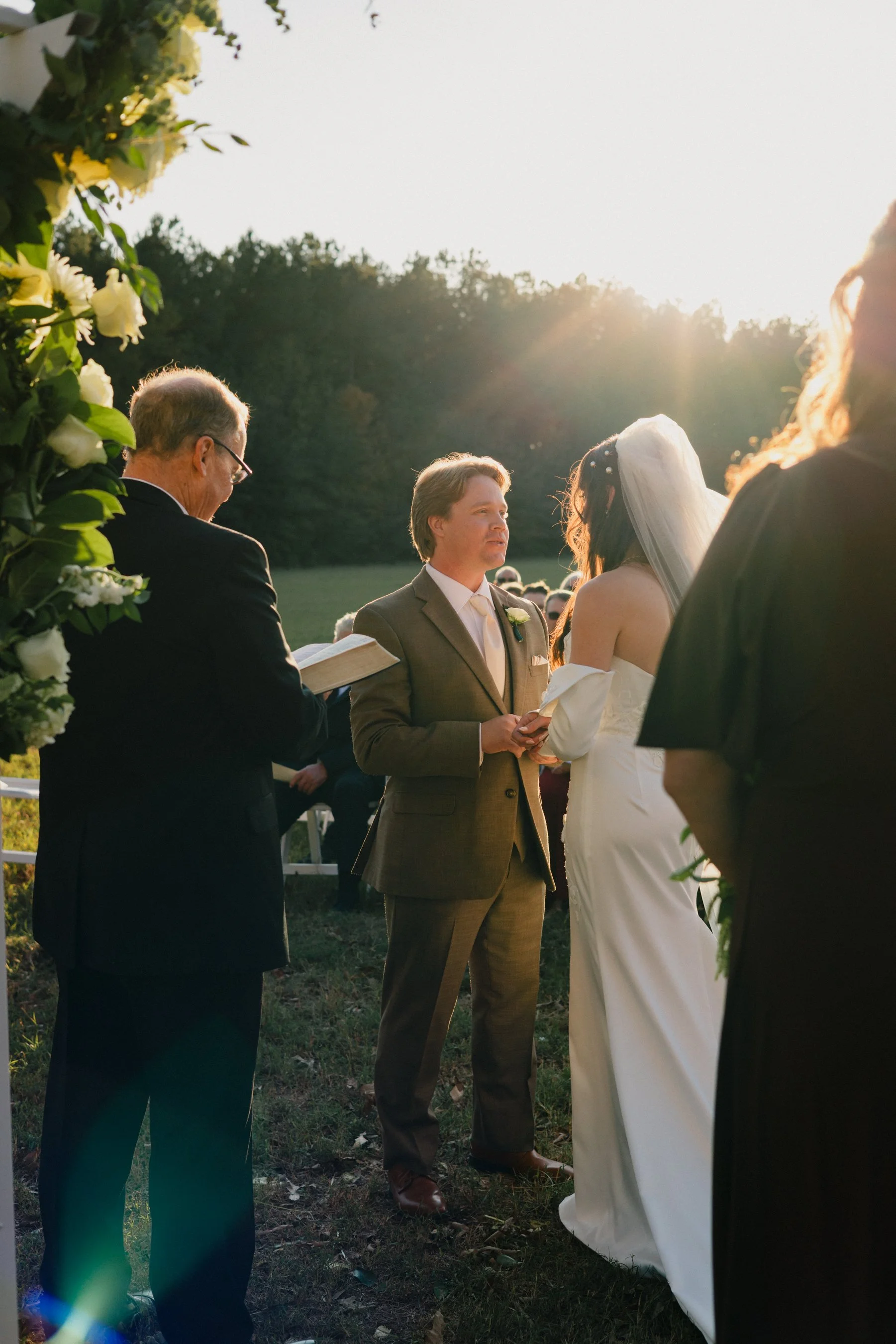 A couple getting married outdoors during sunset, with an officiant reading from a book and a man in a suit holding a paper, framed by floral decorations.