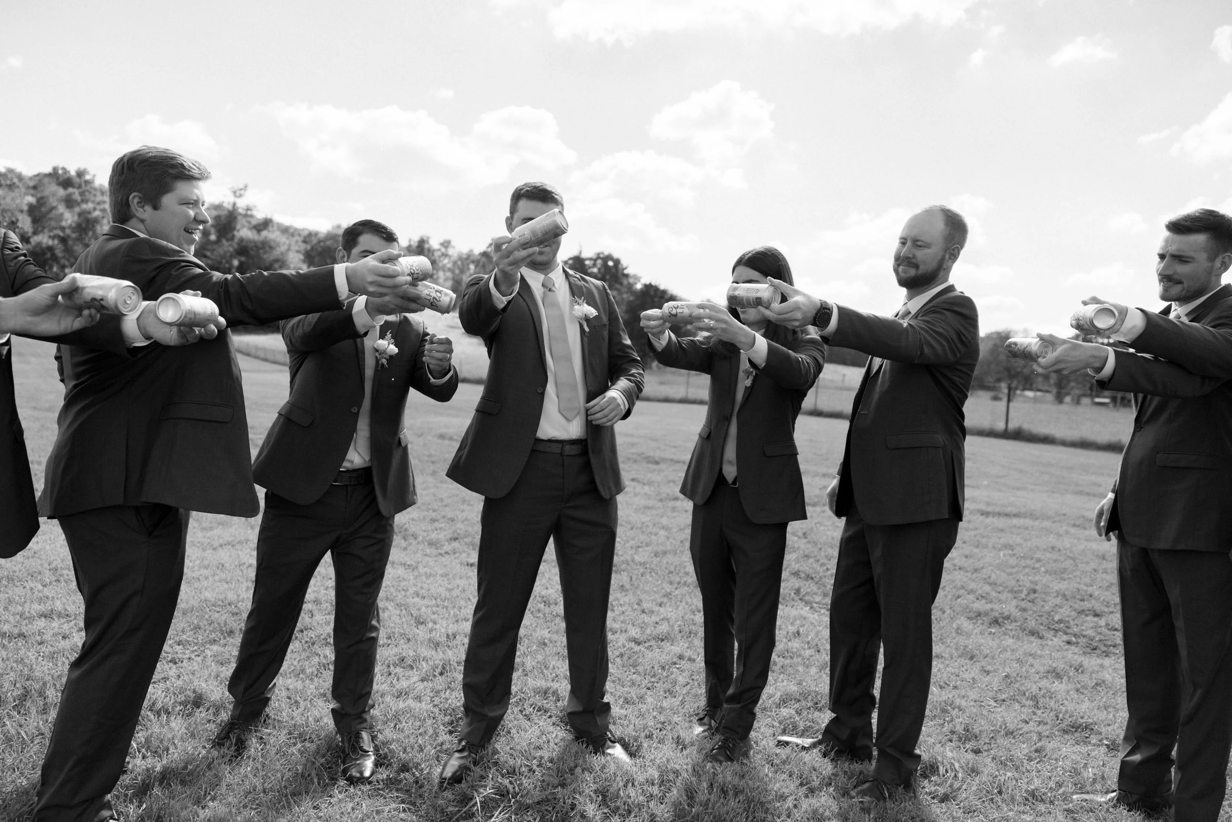 Group of men in suits holding cans in a celebratory toast outdoors on a grassy field.