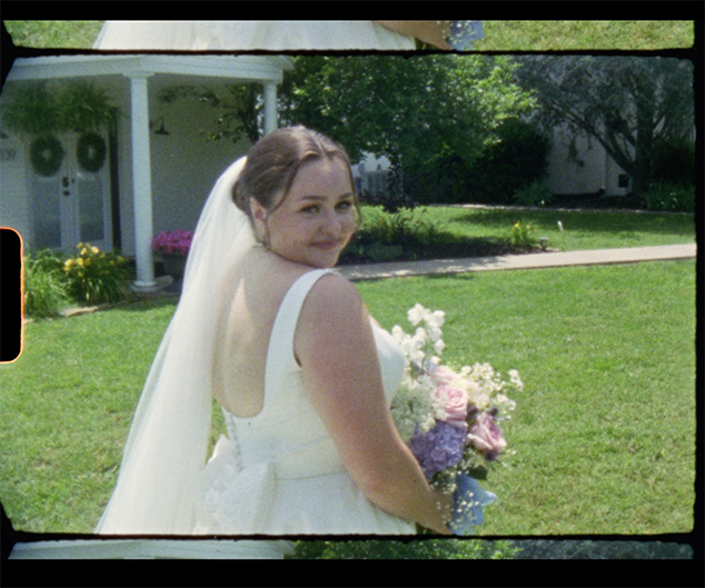 Bride in a white wedding dress holding a bouquet of flowers outdoors on a sunny day, with a house and green lawn in the background.