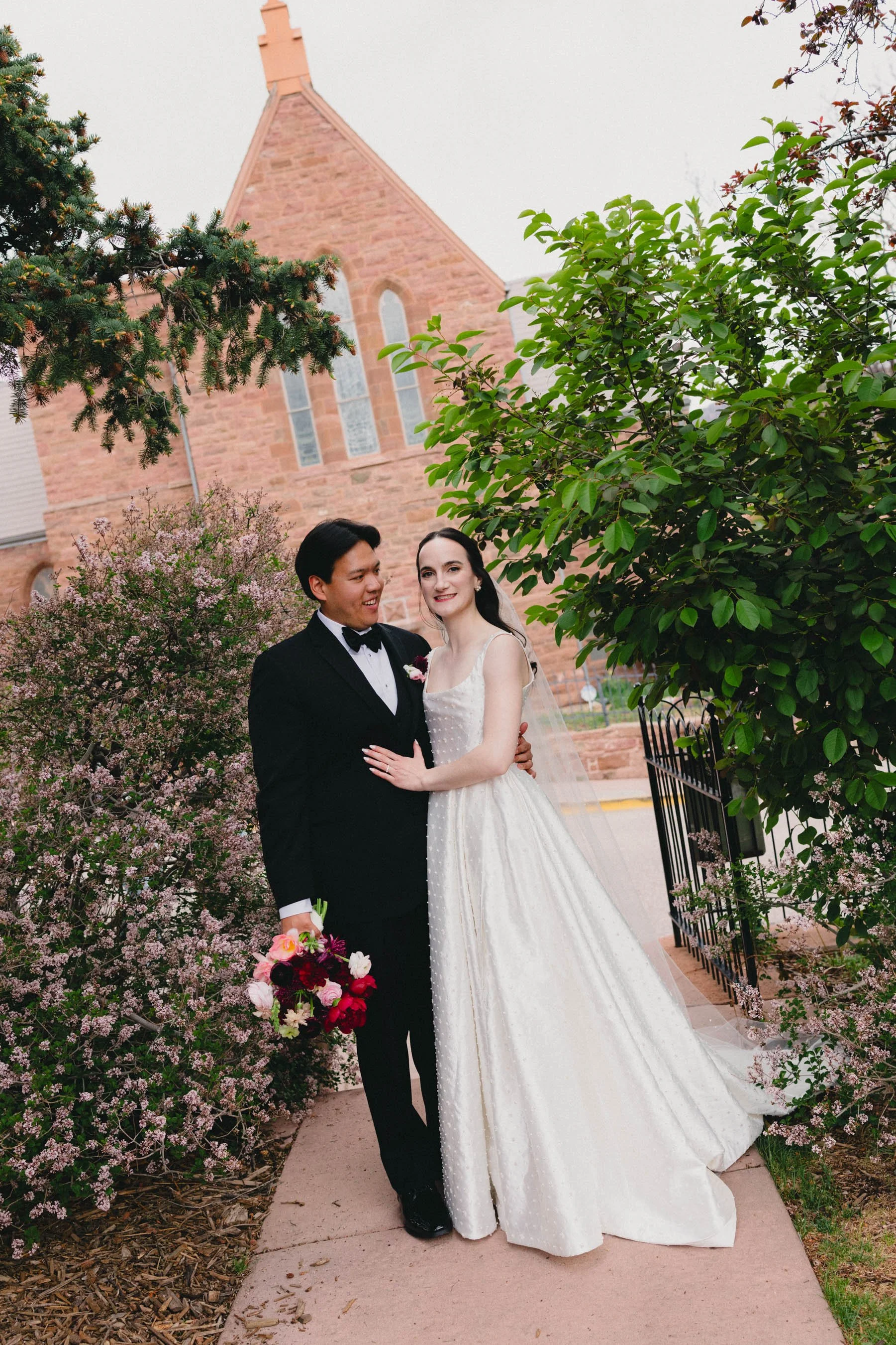 A bride and groom standing outside in front of a Methodist church with large stained glass windows. The bride is wearing a strapless white wedding dress and veil, and the groom is dressed in a black tuxedo with a bowtie. The groom is holding a bouque