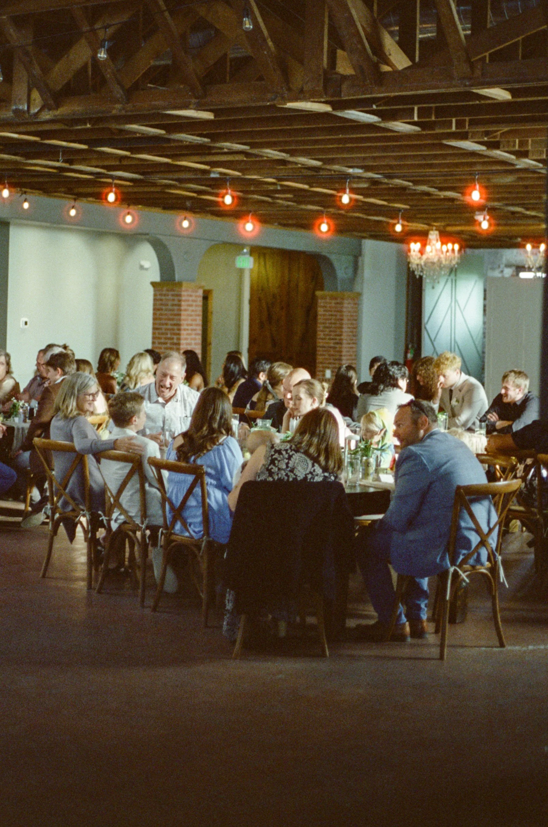 People dining at tables in a restaurant with exposed brick and wooden beams on the ceiling, decorated with string lights and chandeliers.