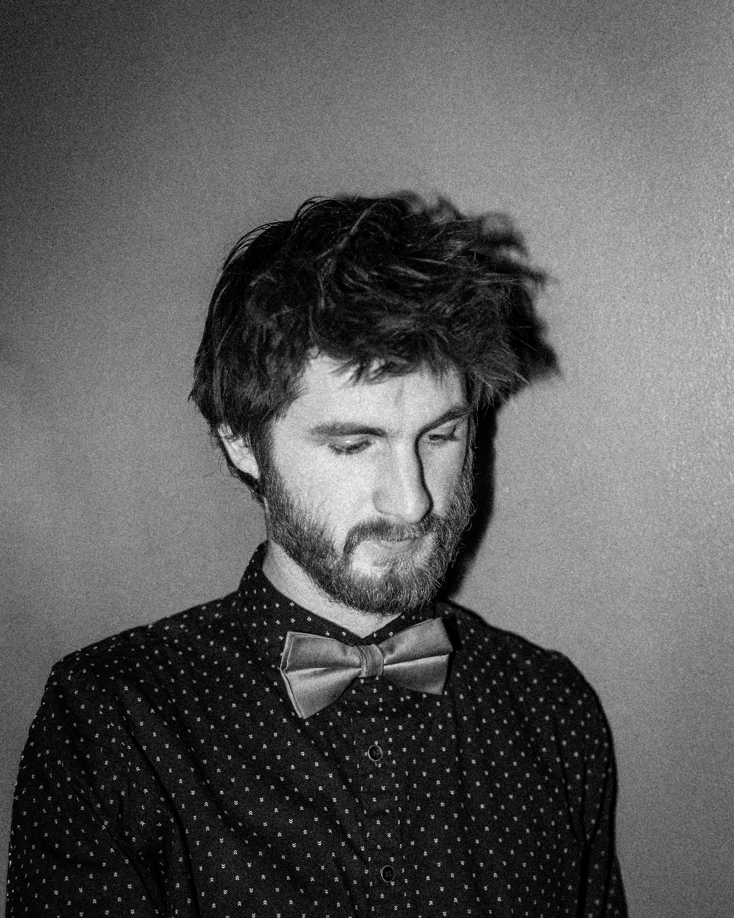 A black and white portrait of a young man with messy hair and a beard, wearing a hoodie.