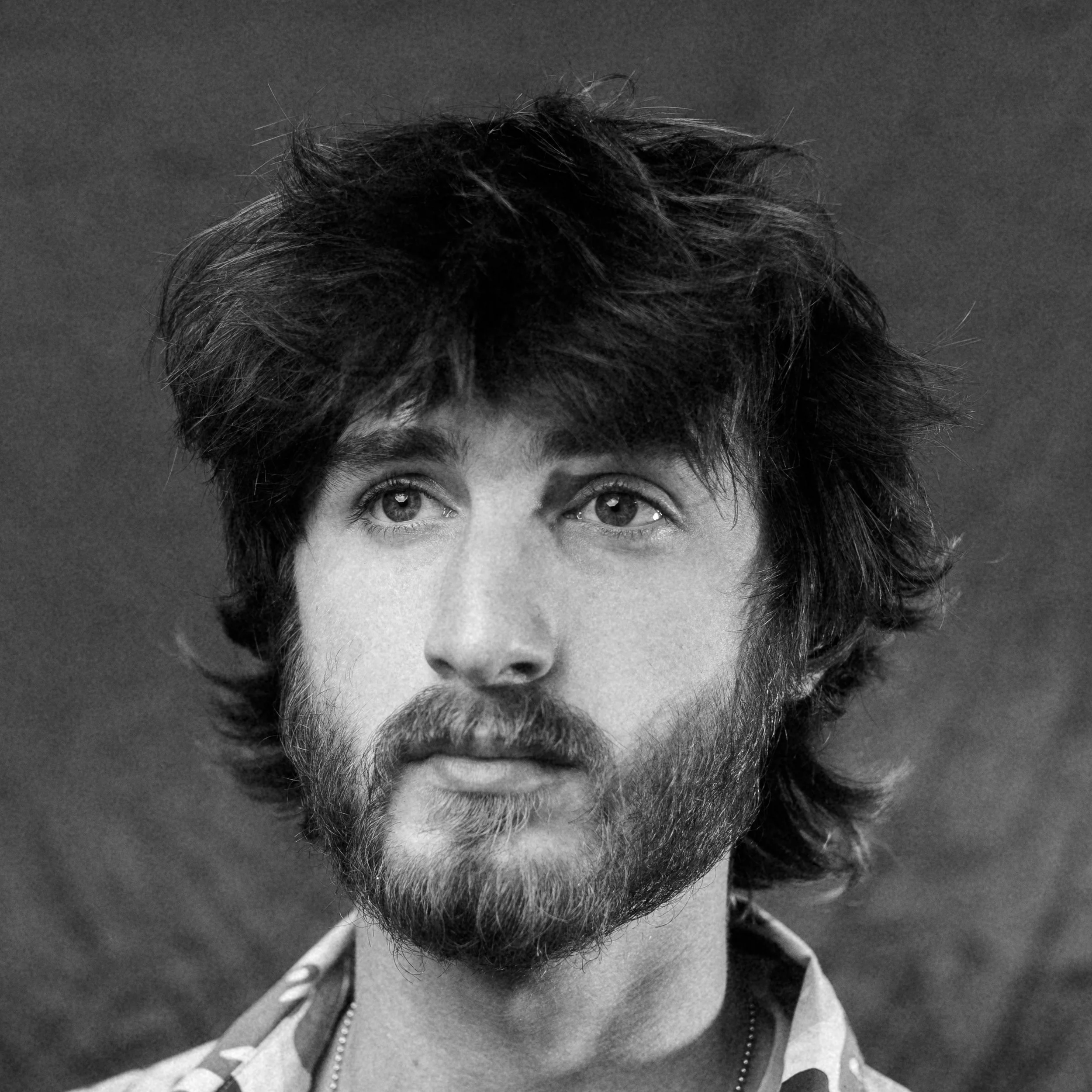 A black and white portrait of a young man with messy hair and a beard, wearing a hoodie.