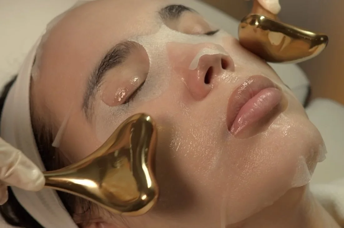 Client receiving facial care with a sheet mask and gold massage tools