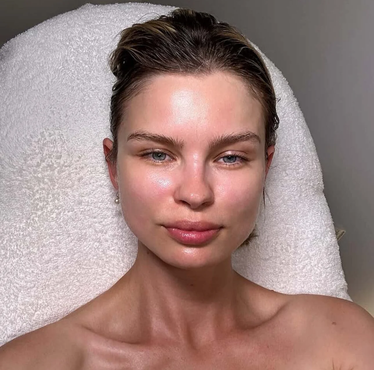 Close-up of a young woman with short, wet hair, blue eyes, and natural makeup, lying on a fluffy white towel against a gray background.