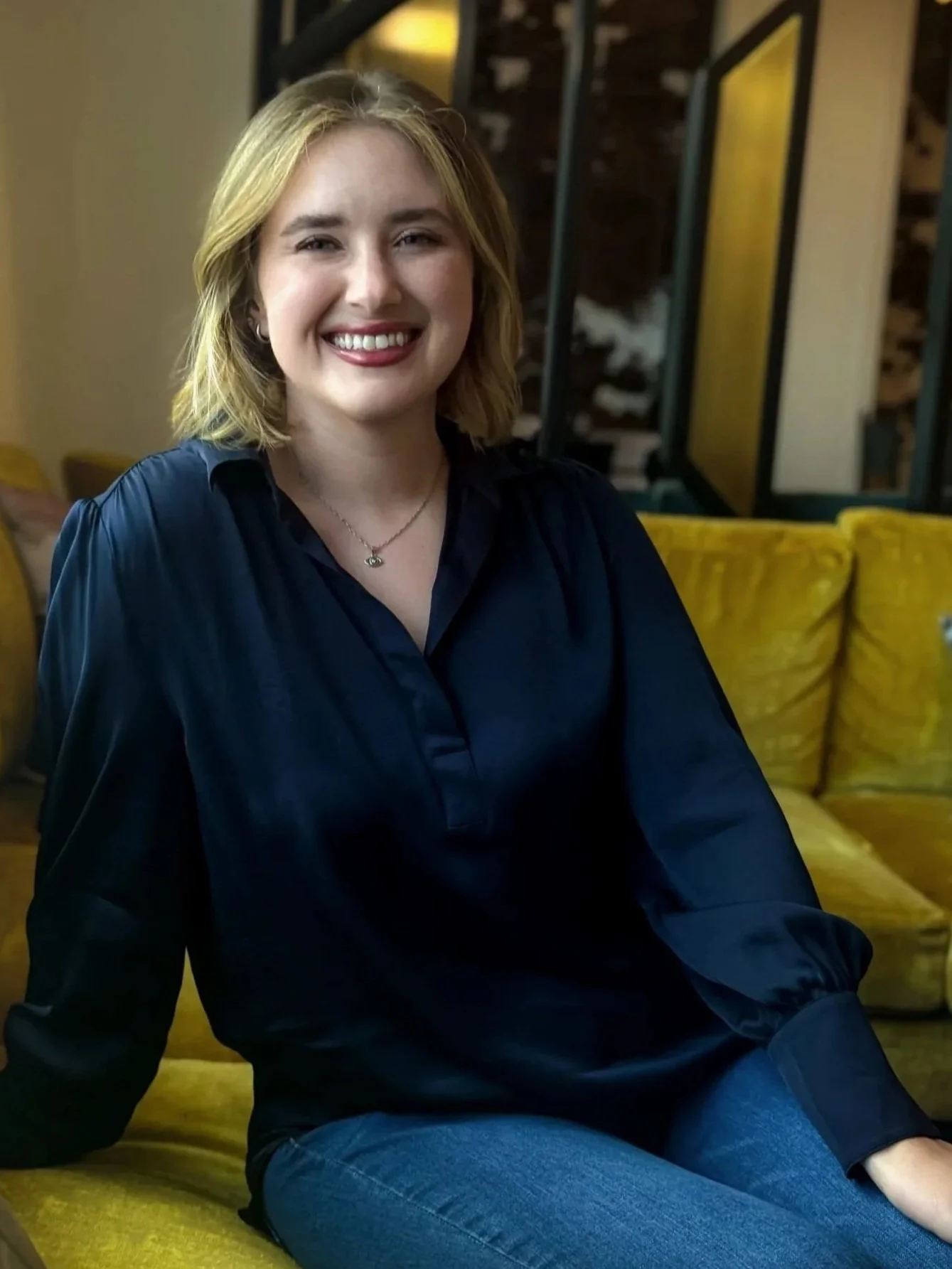 A young woman with shoulder-length blonde hair smiling while sitting on a yellow couch in a cozy, warmly lit room.
